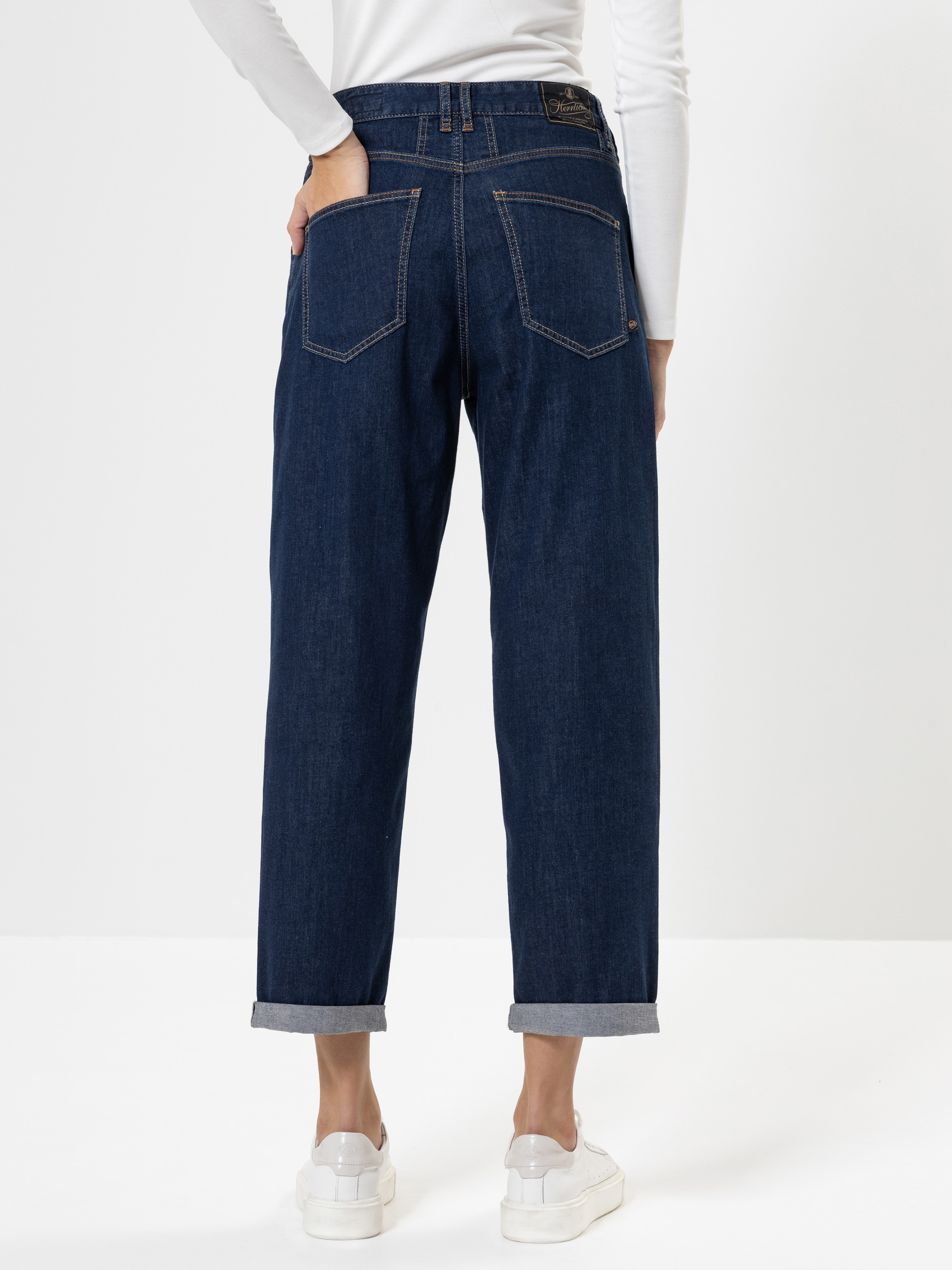 Eine Person, die dunkelblaue Jeans mit Manschetten, ein weißes langärmeliges Oberteil und weiße Turnschuhe trägt, wird von hinten gezeigt, mit einer Hand in der Gesäßtasche, vor einem schlichten weißen Hintergrund stehend.