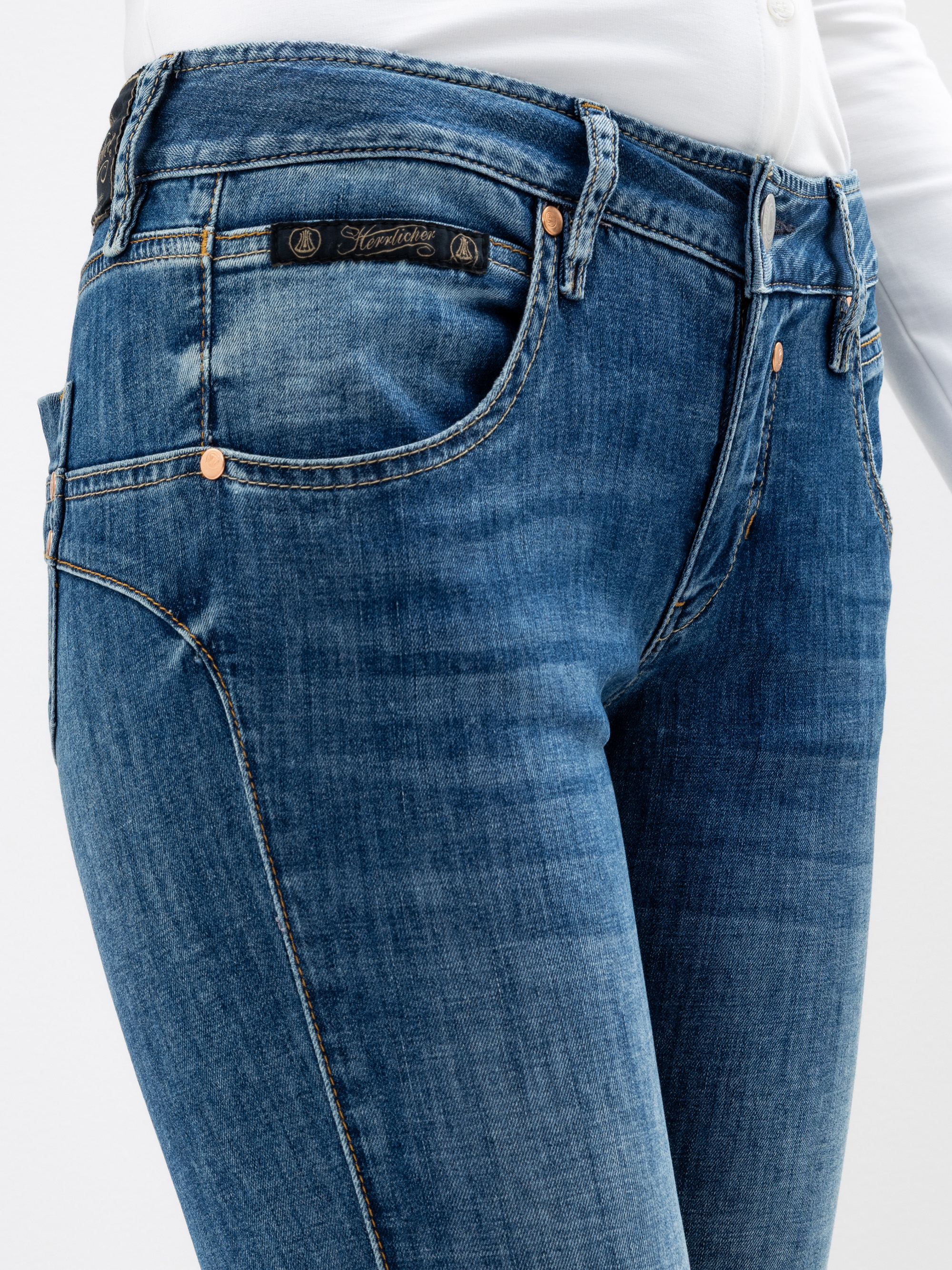 Nahaufnahme einer Person, die eine blaue Jeans und ein weißes Hemd trägt, wobei der Schwerpunkt auf der Taille, den Hüften und den Oberschenkeln liegt. Die Jeans hat kupferne Nieten und ein dunkles Etikett über der rechten Tasche.