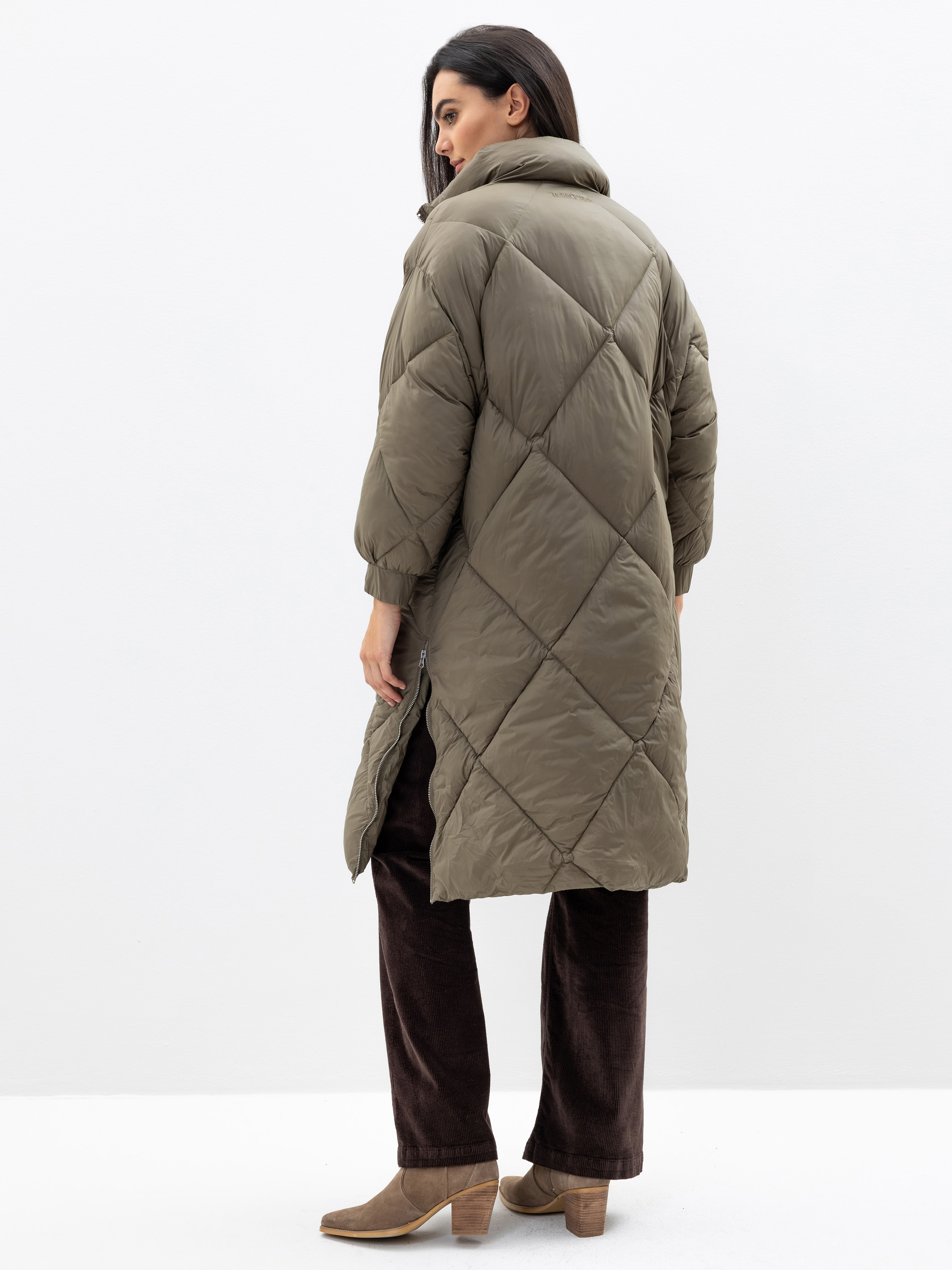 Eine Frau steht mit dem Gesicht zur Wand und trägt einen langen, gesteppten, olivgrünen Puffermantel, eine dunkelbraune Hose und hellbraune, hochhackige Stiefel vor einem schlichten weißen Hintergrund.