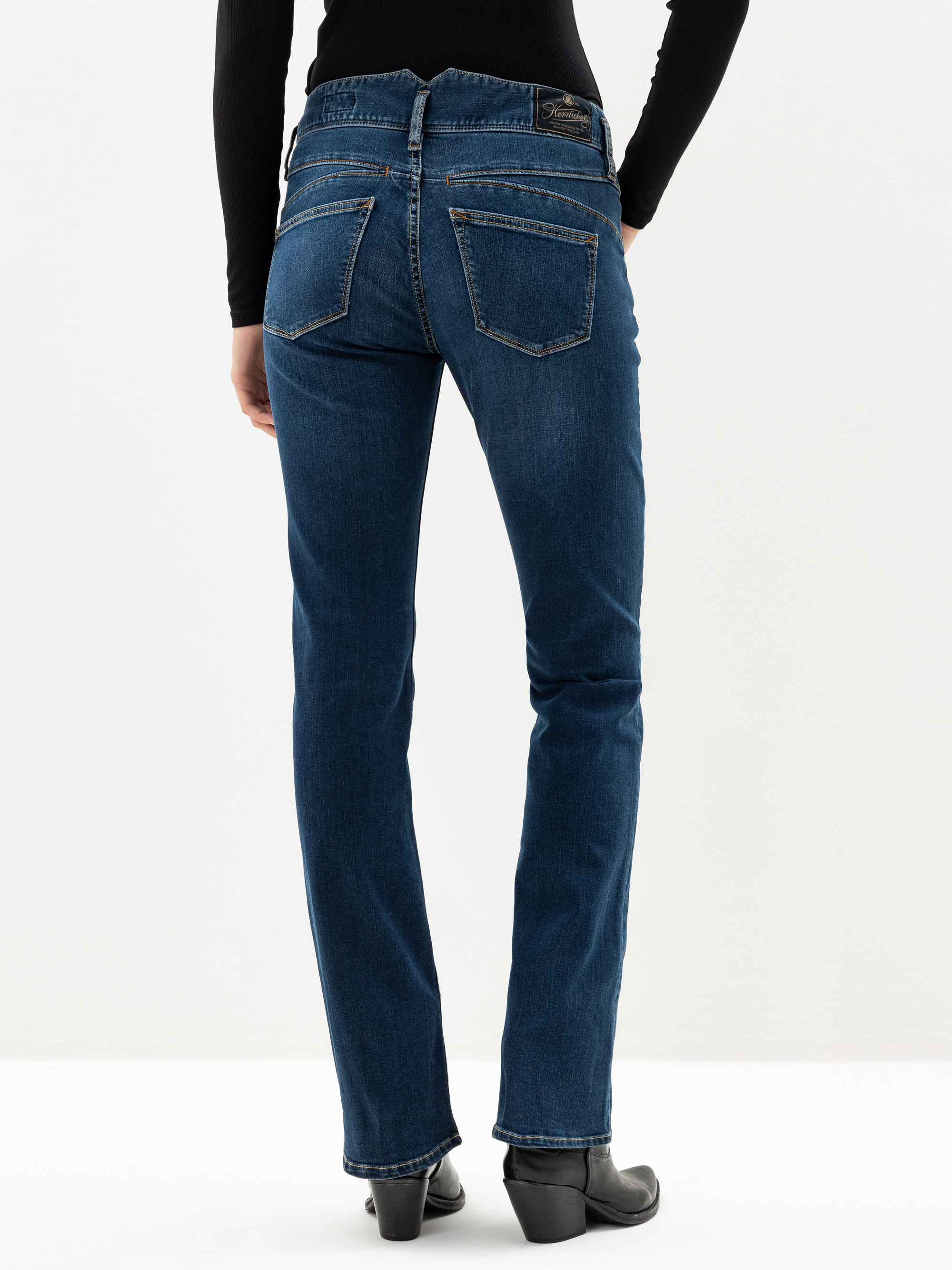 Eine Person, die blaue Jeans mit geradem Bein, schwarze Stiefel mit Absatz und ein schwarzes langärmeliges Oberteil trägt, steht mit dem Rücken zur Kamera vor einem schlichten weißen Hintergrund.