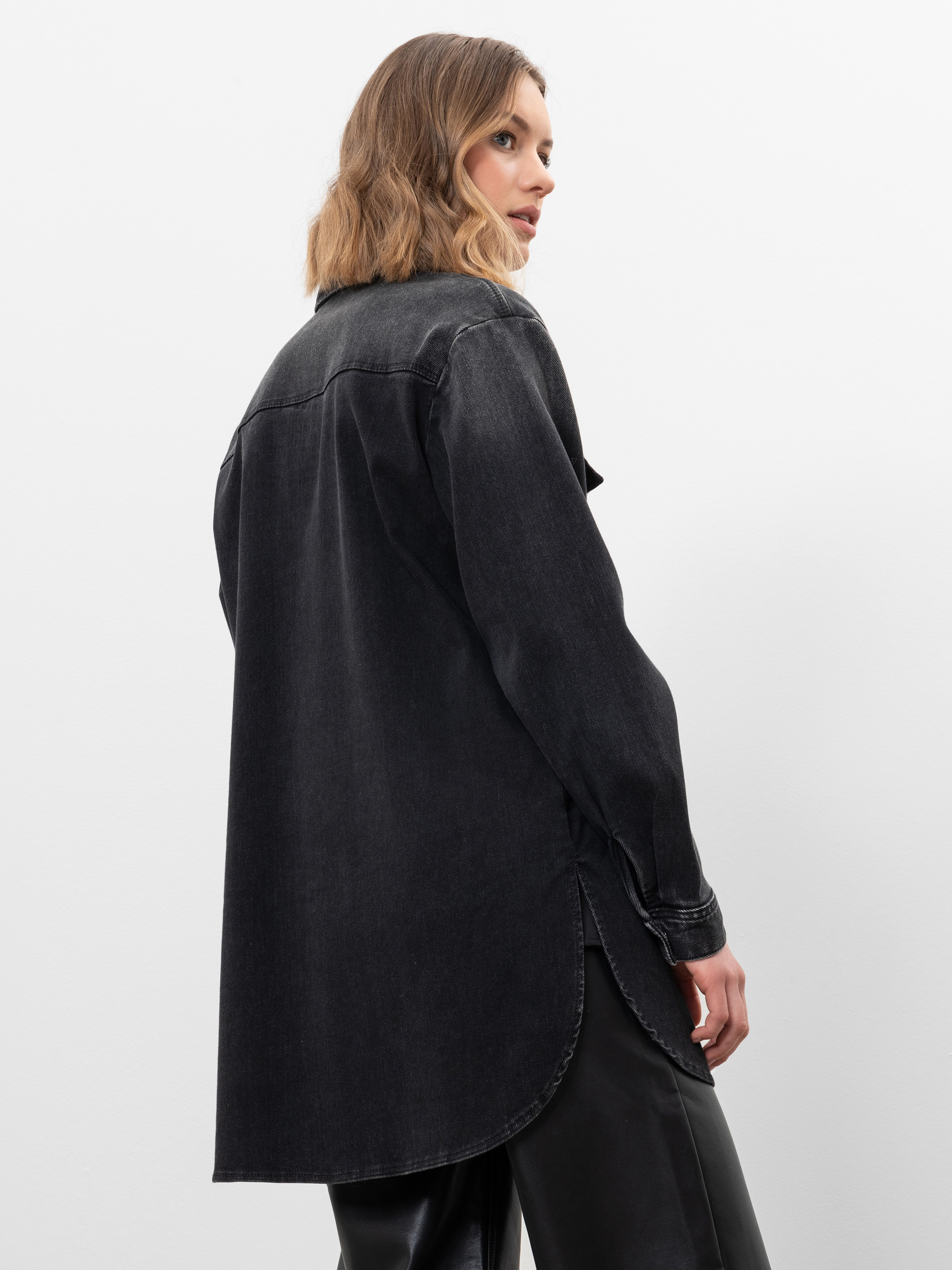 Eine Frau mit schulterlangem hellbraunem Haar trägt ein langes, übergroßes schwarzes Jeanshemd und eine schwarze Hose. Sie steht seitlich und blickt über ihre Schulter vor einem schlichten weißen Hintergrund.