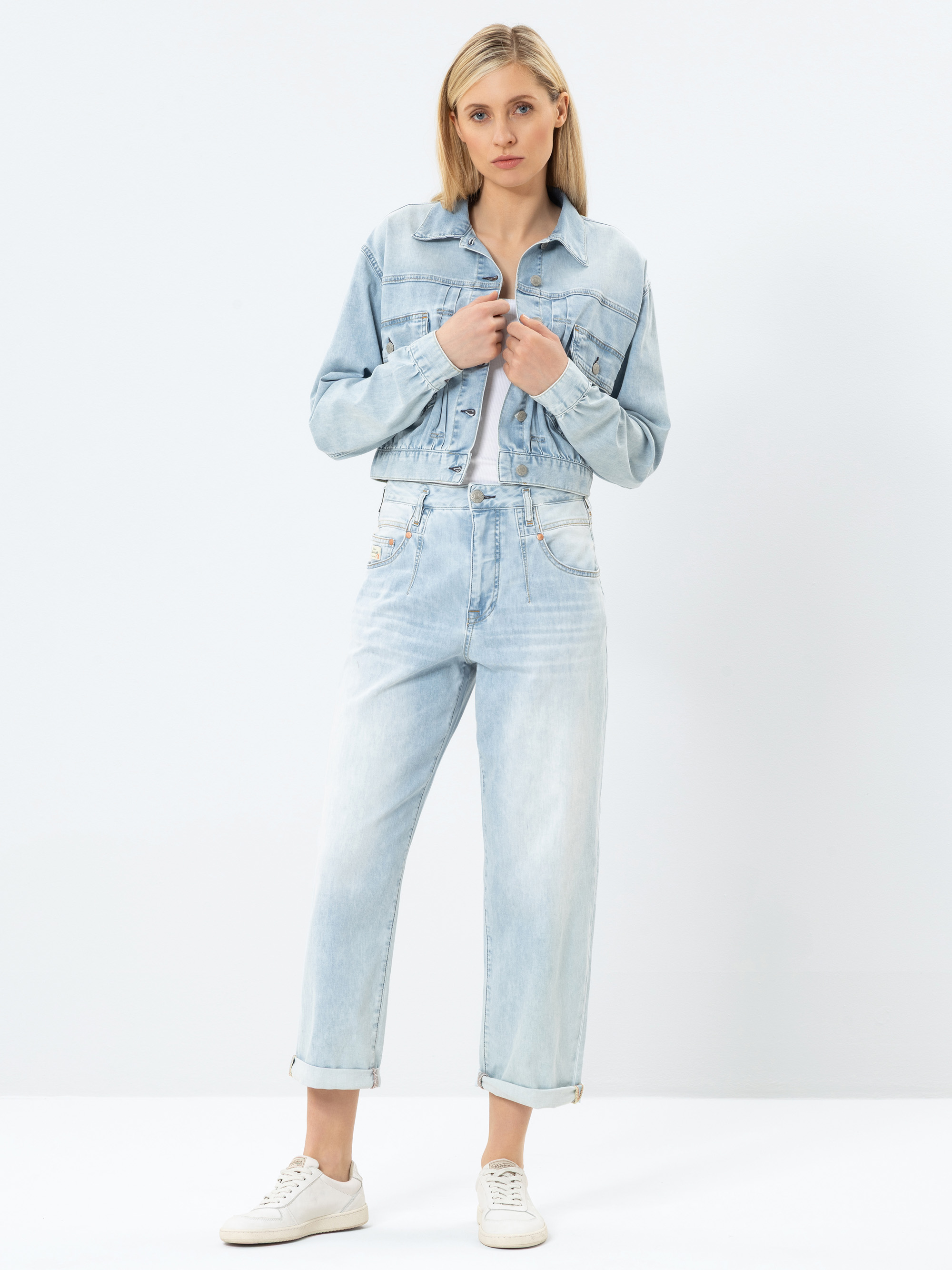 Eine Frau steht vor einem schlichten, hellen Hintergrund. Sie trägt eine hellblaue Jeansjacke und eine dazu passende hochgekrempelte Jeans mit aufgerollten Bündchen, dazu weiße Turnschuhe. Sie hat glattes blondes Haar und einen neutralen Gesichtsausdru
