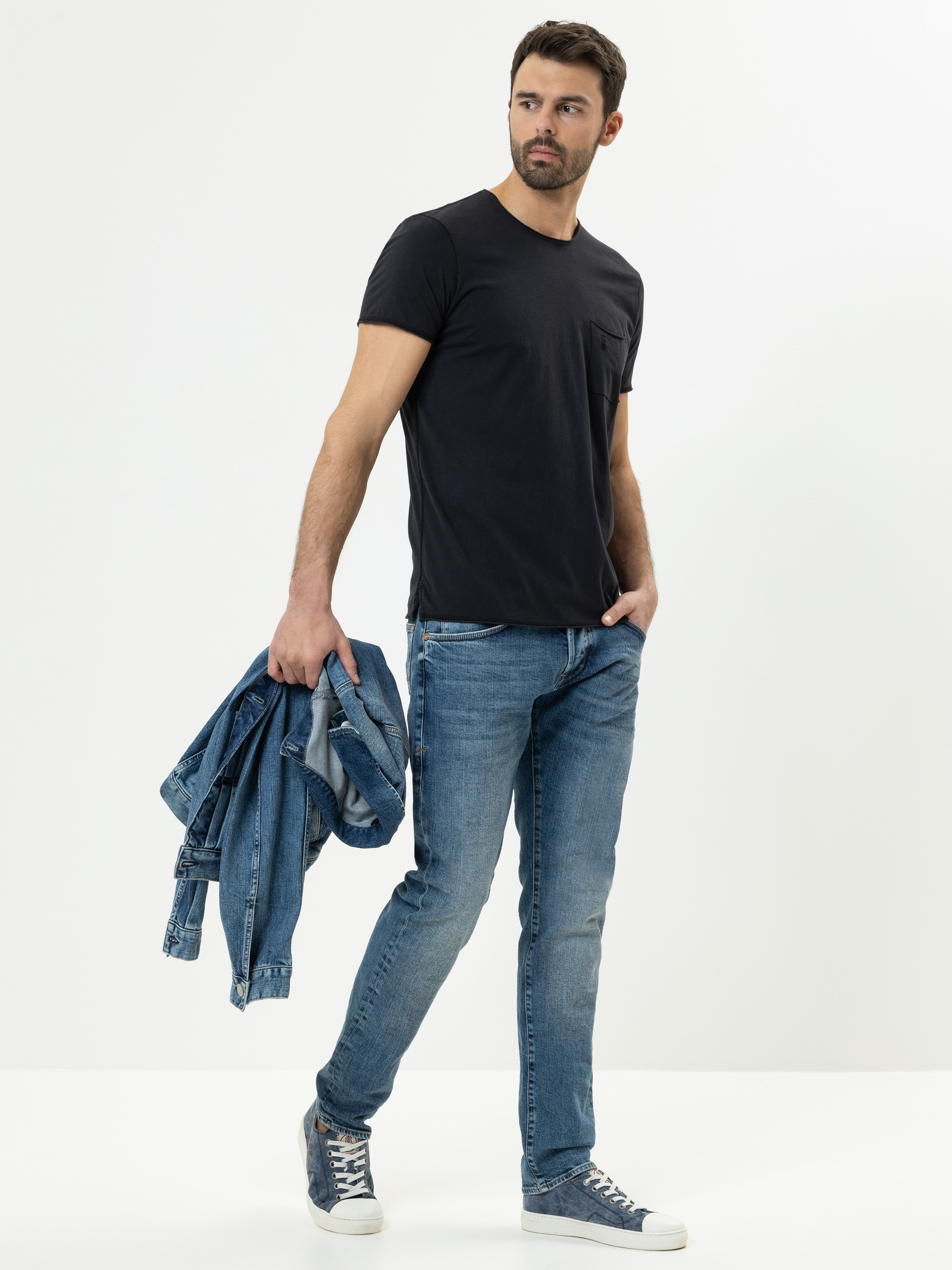Ein Mann, der ein schwarzes T-Shirt, blaue Jeans und blaue Turnschuhe trägt, steht vor einem weißen Hintergrund und hält eine blaue Jeansjacke in der linken Hand, während er nach rechts schaut.