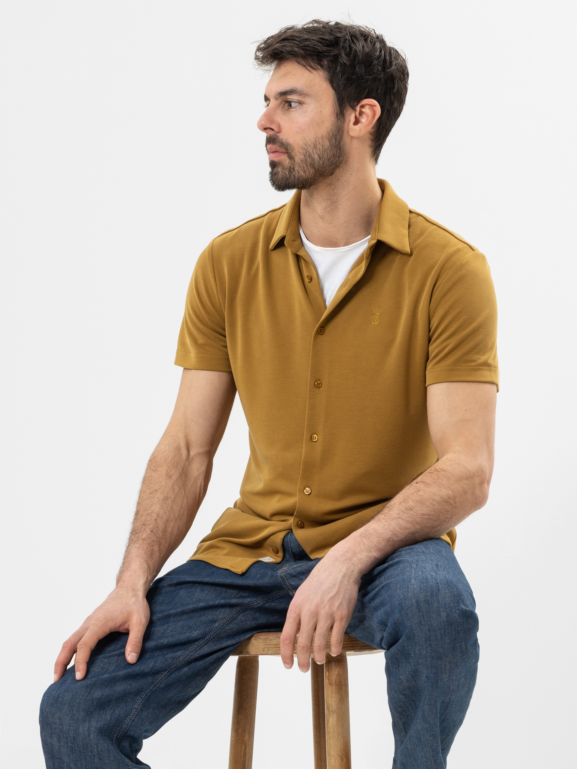 Ein Mann mit dunklem Haar und Bart sitzt auf einem Holzschemel und trägt ein senffarbenes, kurzärmeliges Button-up-Hemd über einem weißen T-Shirt und blaue Jeans. Er blickt leicht nach links vor einem schlichten weißen Hintergrund.