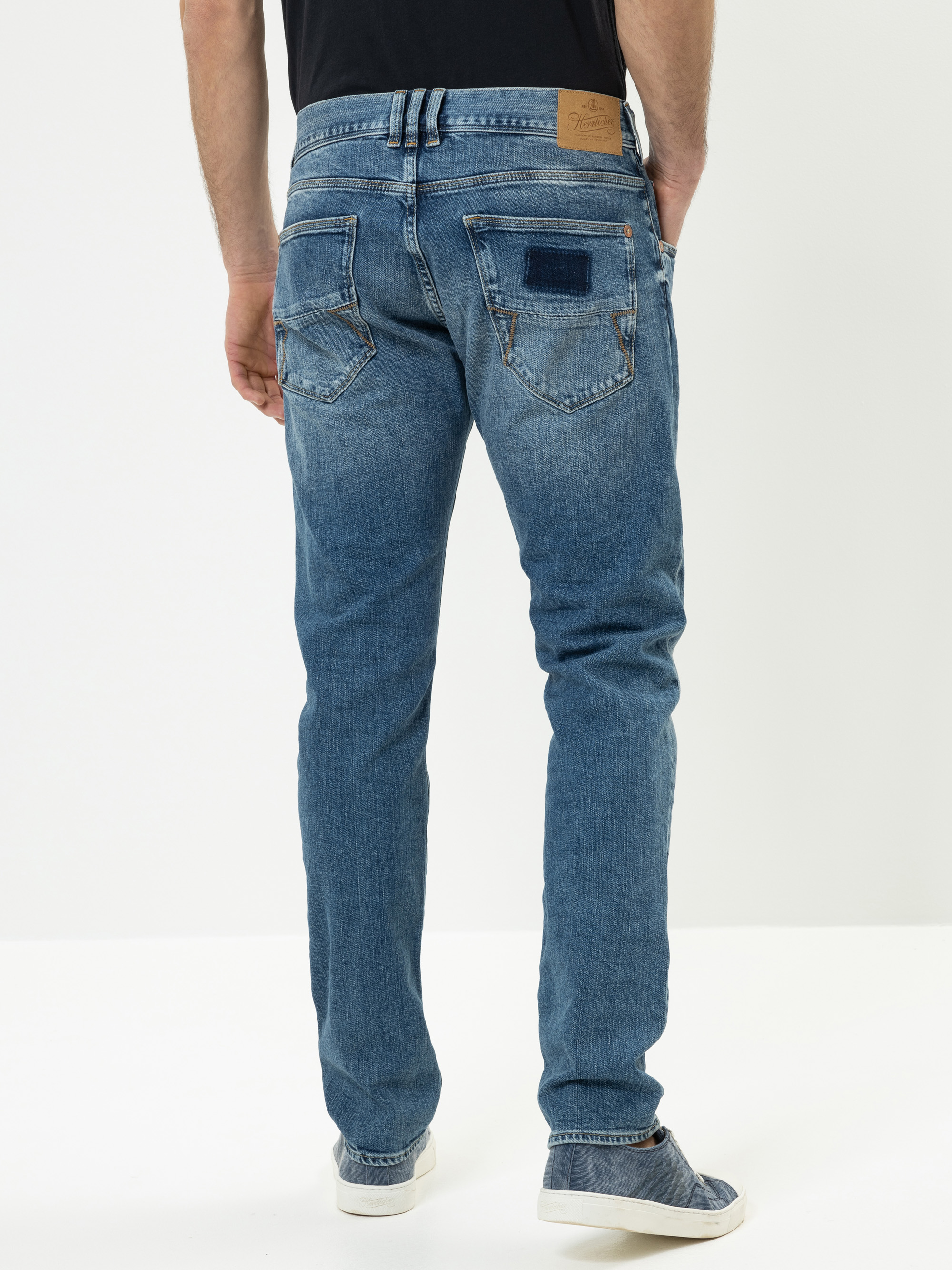 Eine Person, die eine blaue Jeans, ein schwarzes Hemd und weiße Schuhe trägt, steht mit dem Gesicht nach hinten und zeigt die Rückenansicht der Hose vor einem hellen Hintergrund.