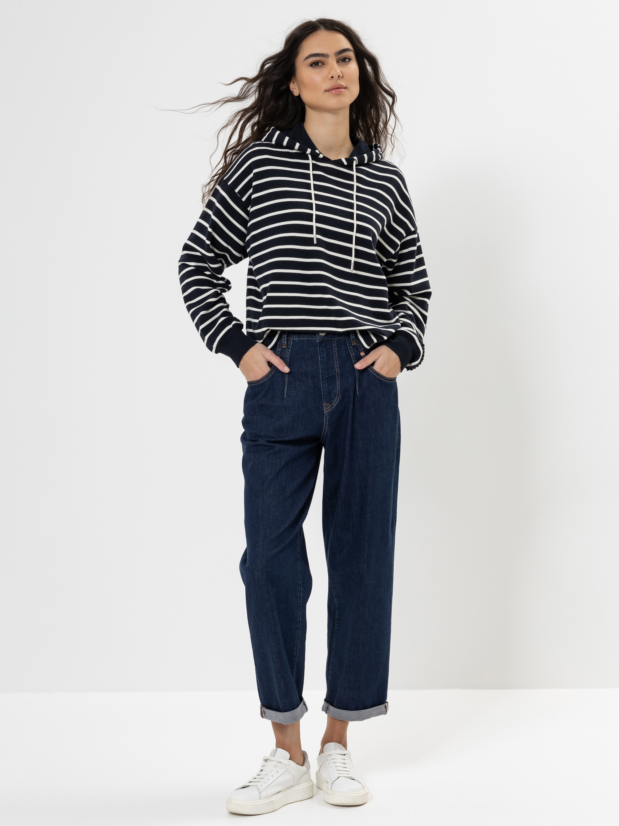 Eine Frau steht vor einem weißen Hintergrund, trägt einen schwarz-weiß gestreiften Kapuzenpullover, dunkelblaue Jeans mit Bündchen und weiße Turnschuhe, die Hände in den Taschen und das Haar offen.