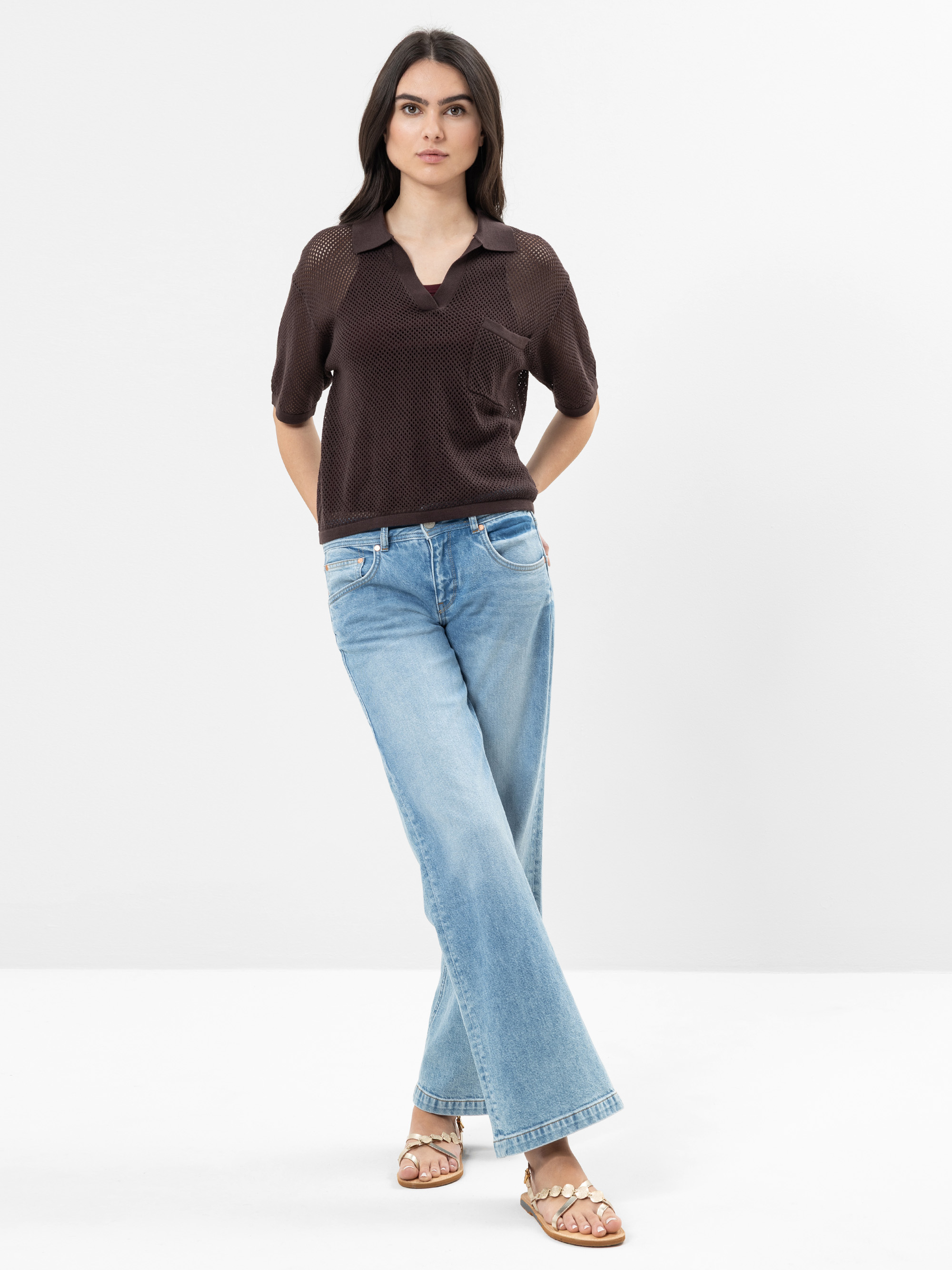 Eine Frau steht vor einem schlichten weißen Hintergrund und trägt ein dunkelbraunes kurzärmeliges Oberteil, hellblaue Jeans mit weitem Bein und hellbraune Sandalen. Sie hat langes, dunkles Haar und einen neutralen Gesichtsausdruck, die Arme sind entspa