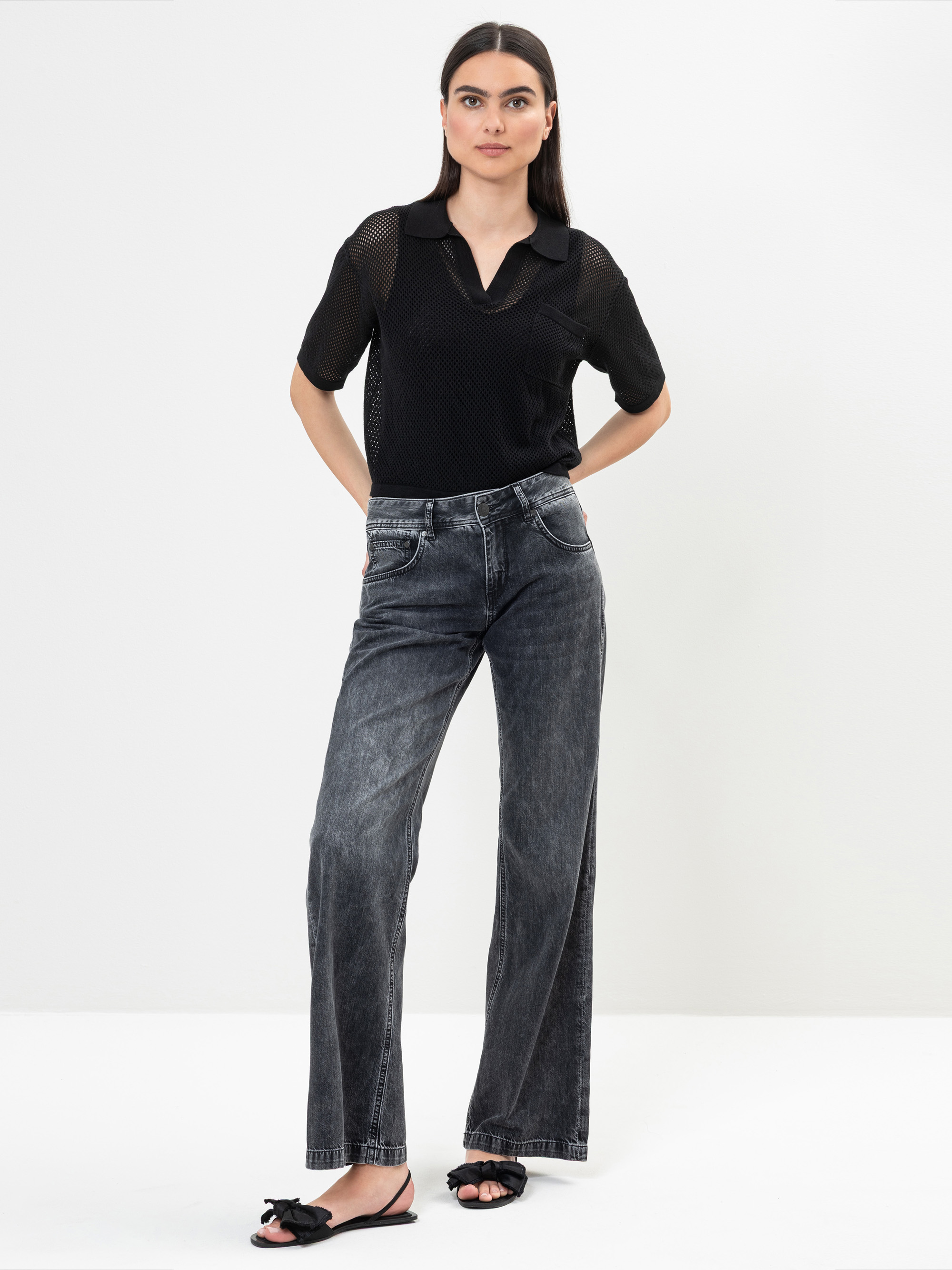 Eine Frau mit langen dunklen Haaren steht vor einem weißen Hintergrund. Sie trägt ein schwarzes kurzärmeliges Hemd, verblichene schwarze Jeans mit weitem Bein und schwarze Sandalen mit Schleifen. Sie hat einen neutralen Gesichtsausdruck und die Hände