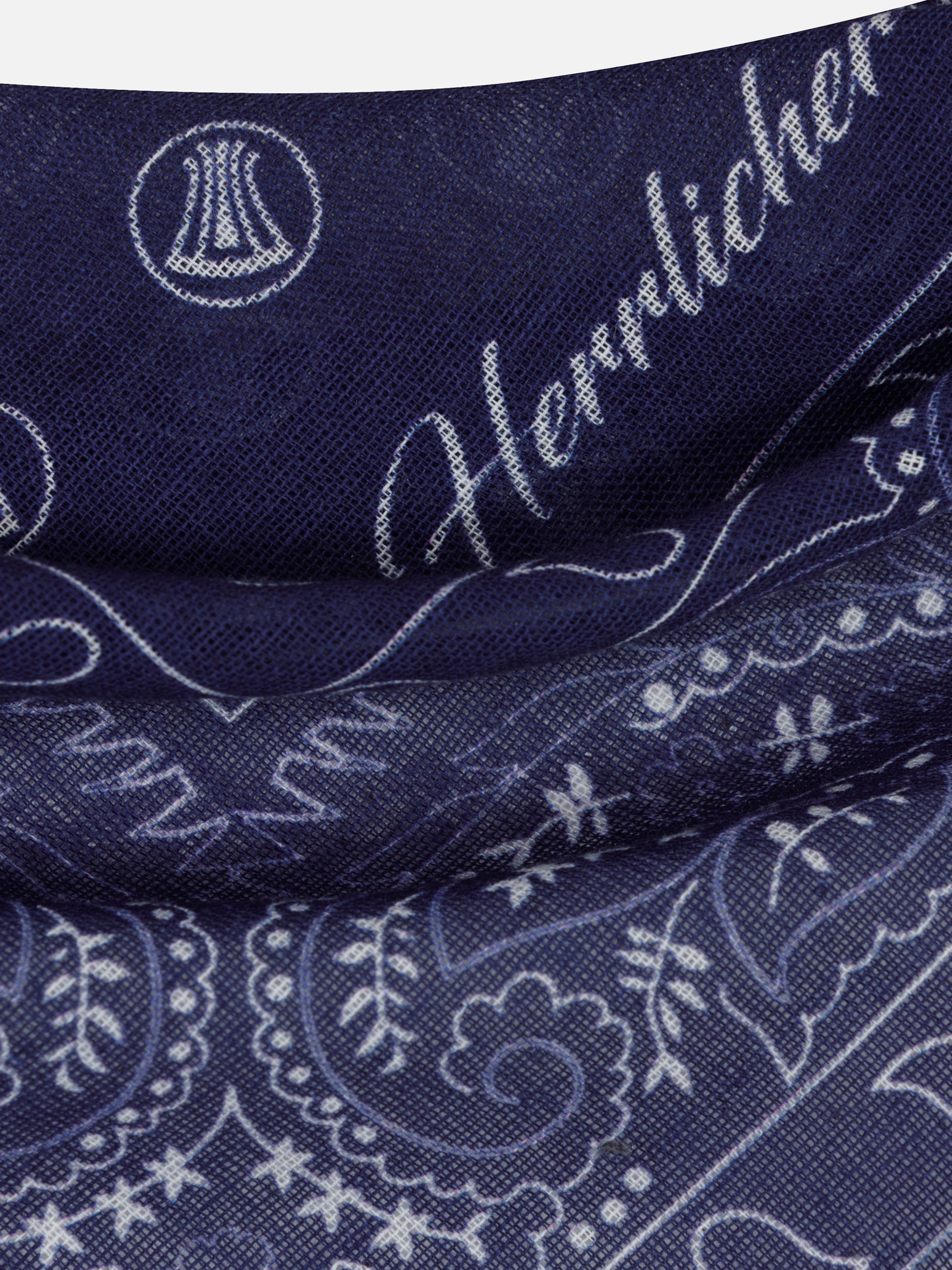 Nahaufnahme eines dunkelblauen Stoffes mit weißen Paisley- und Blumenmustern, darunter ein kursives Wort Herrlicher und kreisförmige, eingewebte Emblemmotive.