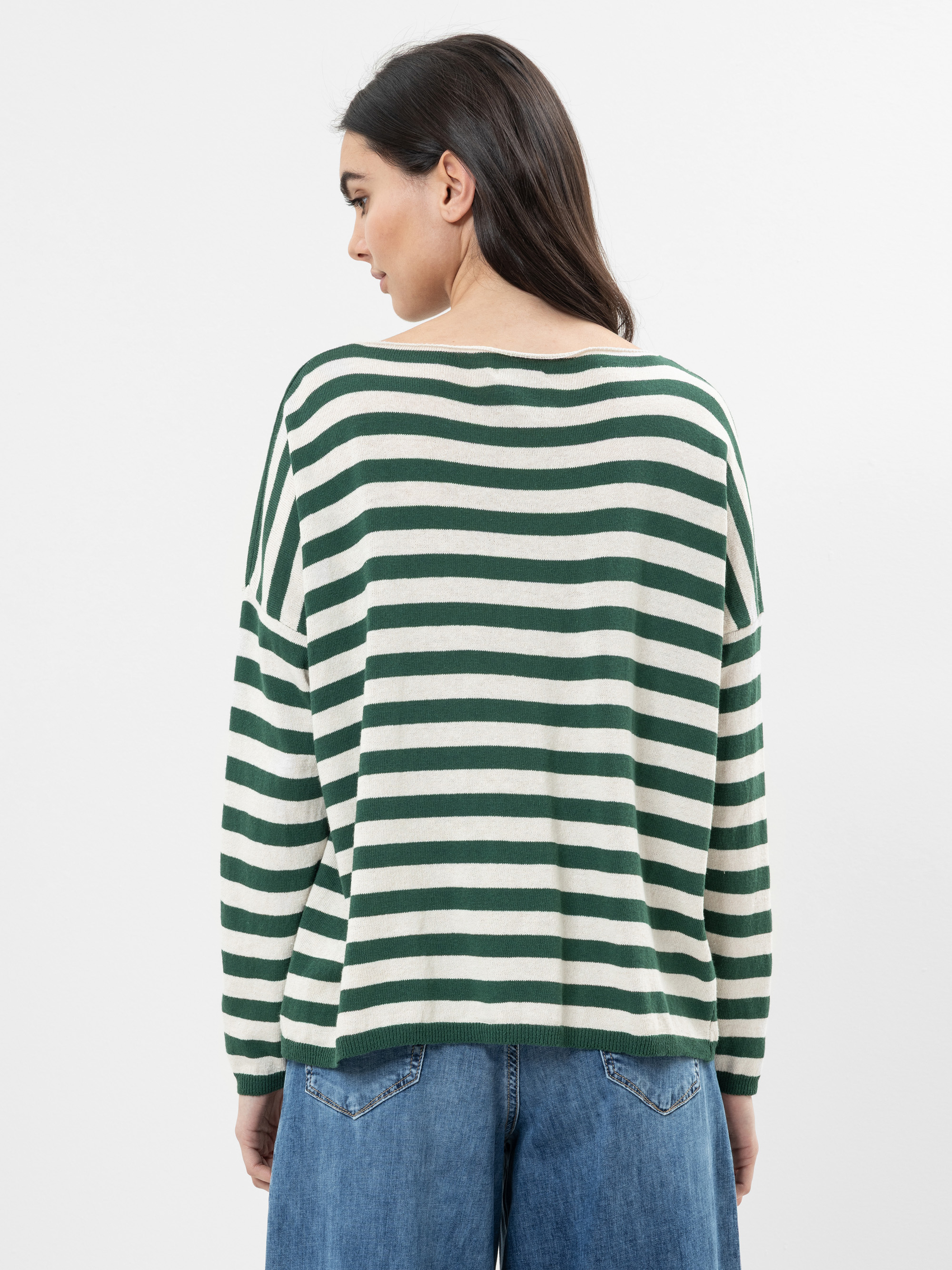 Eine Frau mit langen dunklen Haaren, von hinten gesehen, trägt einen locker sitzenden, grün-weiß horizontal gestreiften Pullover und blaue Jeans vor einem schlichten weißen Hintergrund.