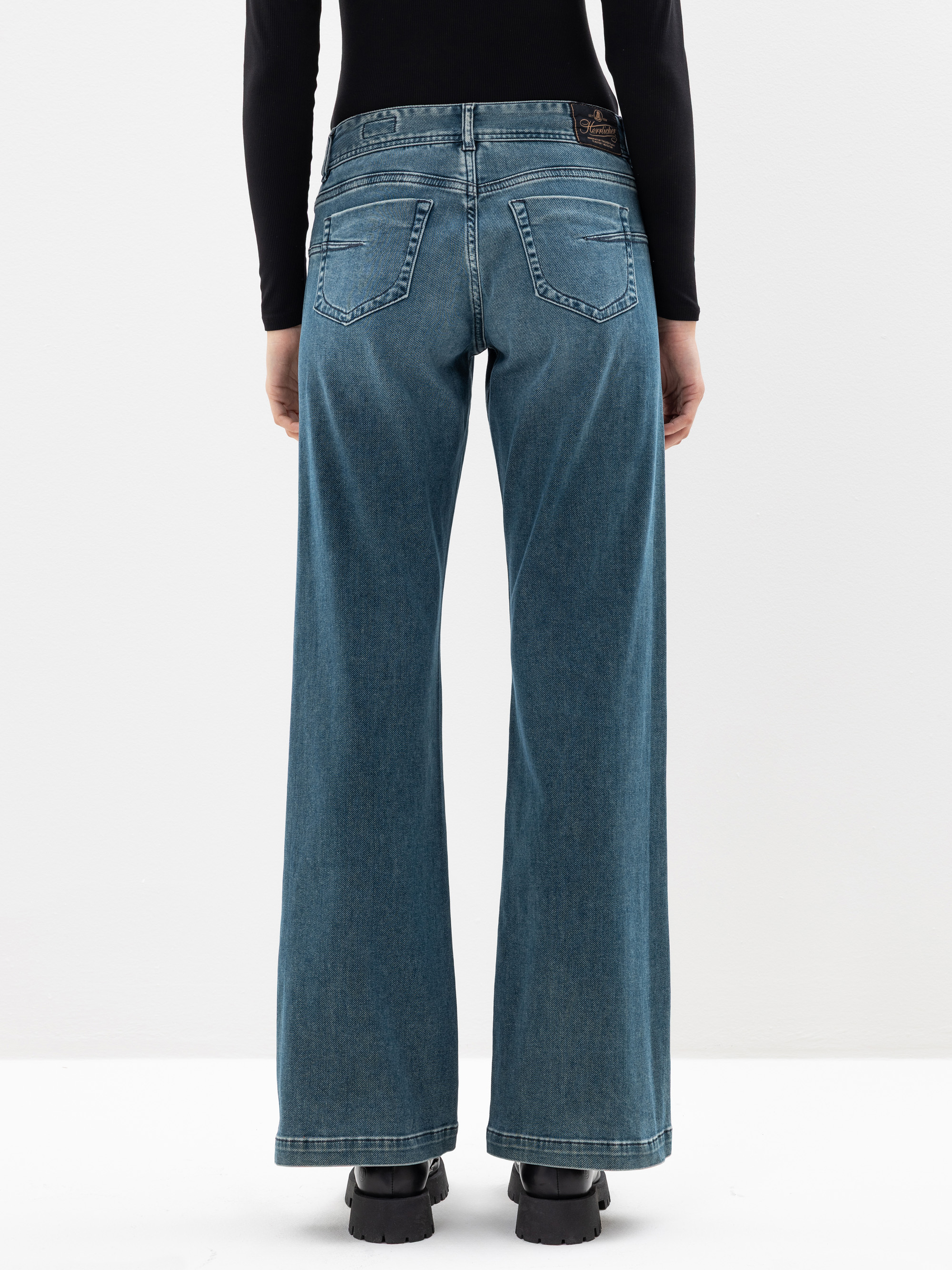 Person, die eine blaue Jeans mit hoher Taille und weitem Bein und ein schwarzes langärmeliges Oberteil trägt, von hinten gesehen. Die Jeans hat zwei Taschen und ist dezent ausgeblichen. Die Person steht vor einem schlichten weißen Hintergrund.