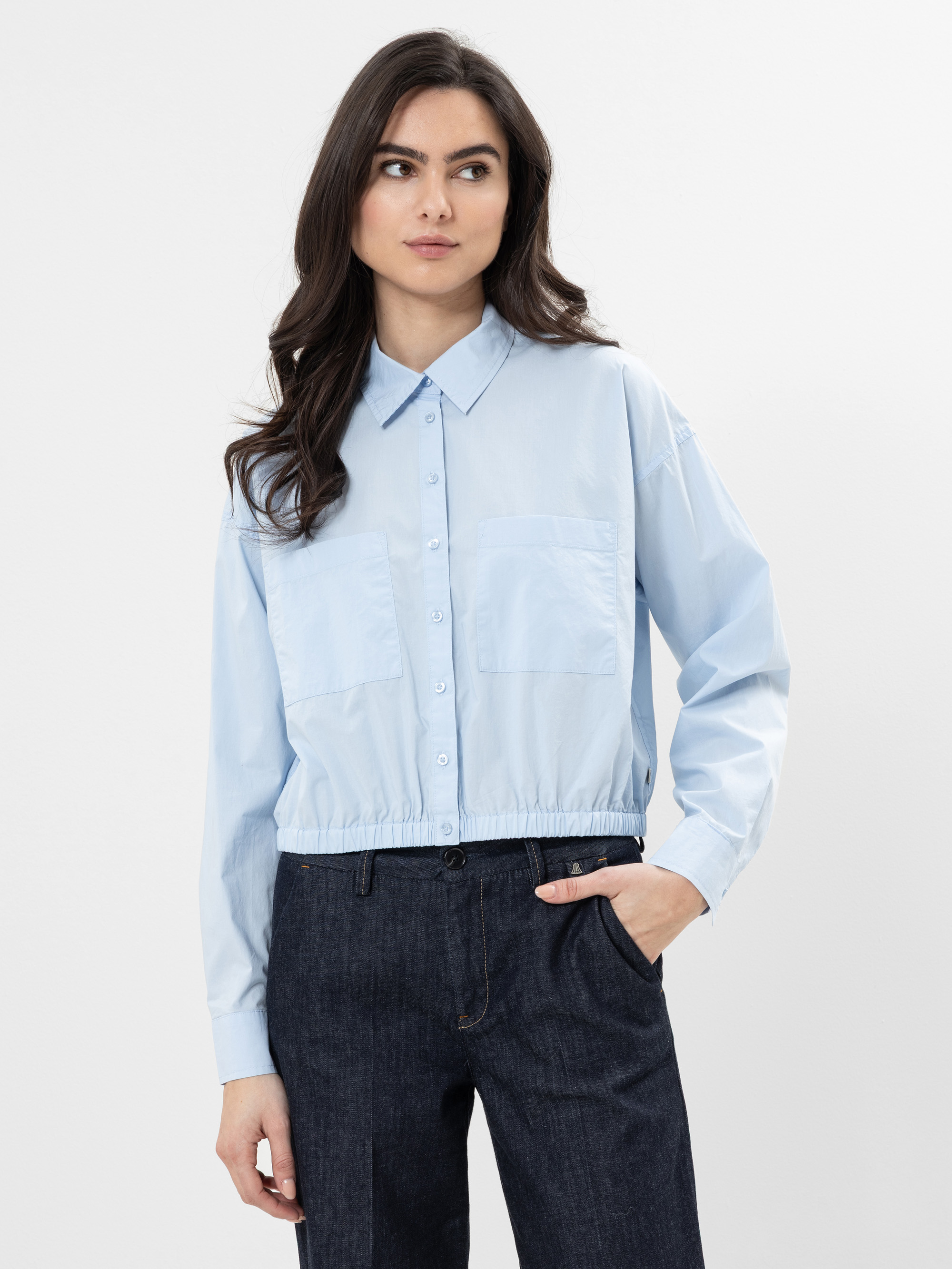 Eine Frau mit langen dunklen Haaren trägt ein hellblaues, geknöpftes Hemd mit zwei Brusttaschen und dunkle Jeans. Sie steht vor einem schlichten weißen Hintergrund und hat eine Hand in der Tasche.