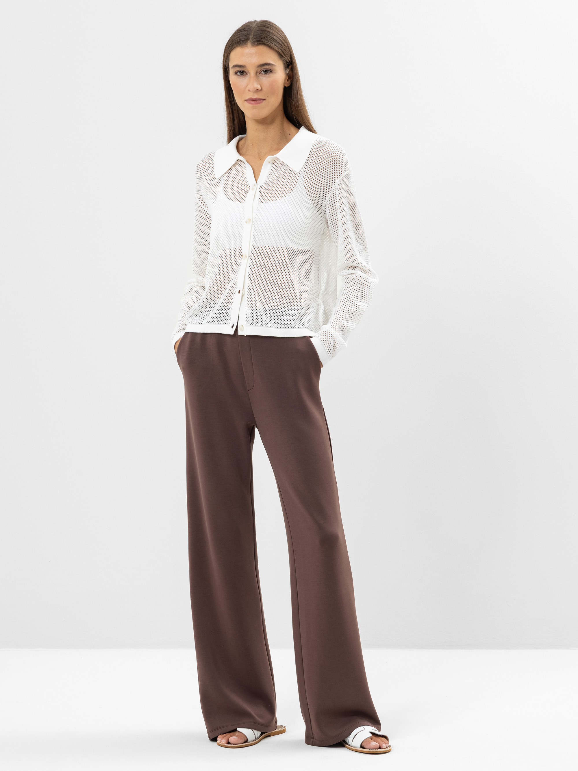 Eine Frau steht vor einem schlichten weißen Hintergrund. Sie trägt ein durchsichtiges weißes Button-up-Hemd über einem weißen Oberteil, weite braune Hosen und weiße Sandalen. Sie hat langes, glattes braunes Haar und ihre Hände stecken in den Tasche