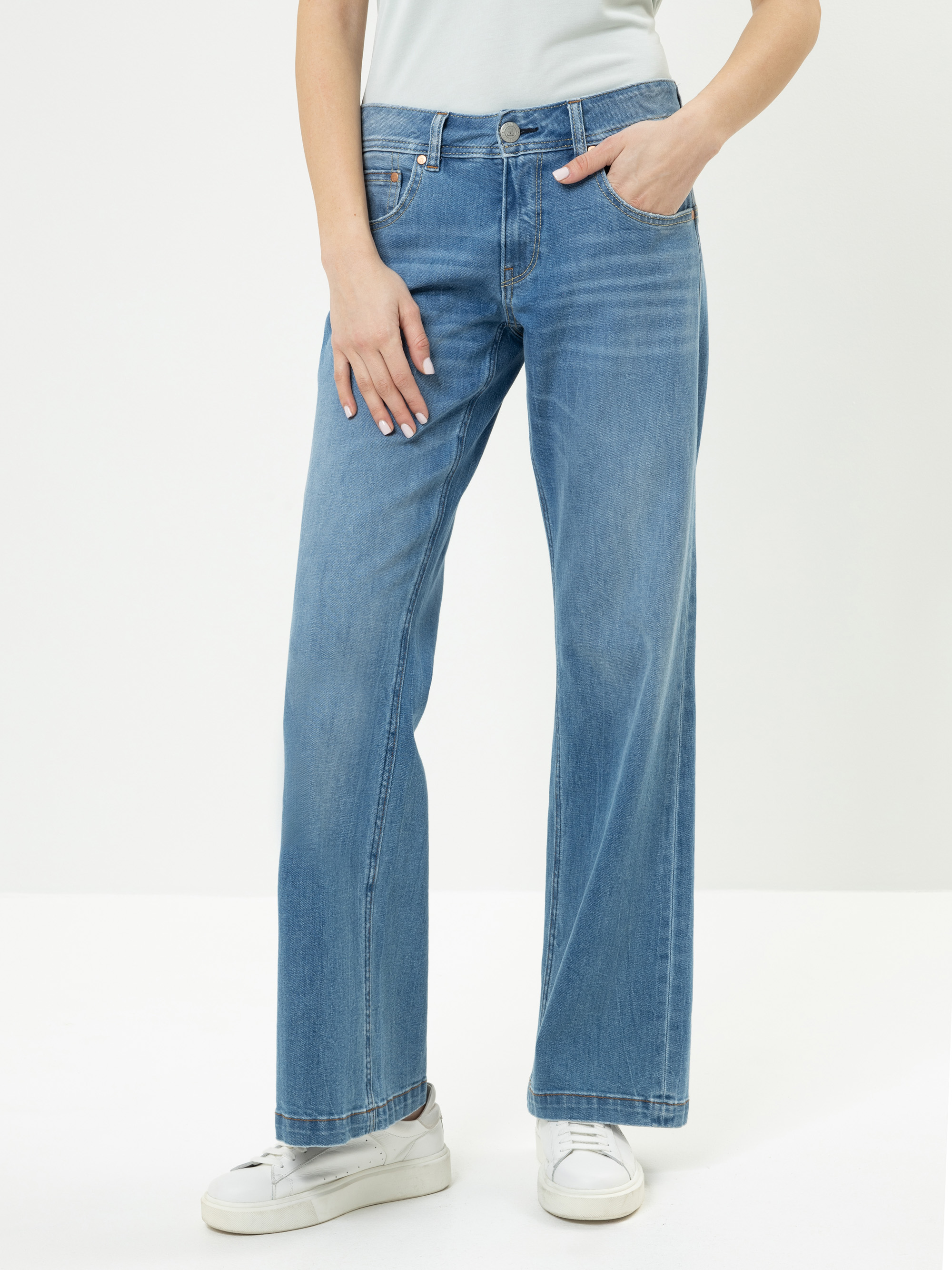 Eine Person in hellblauen, weit geschnittenen Jeans, einem hellen Oberteil und weißen Turnschuhen steht mit einer Hand in der Tasche vor einem schlichten, hellen Hintergrund.