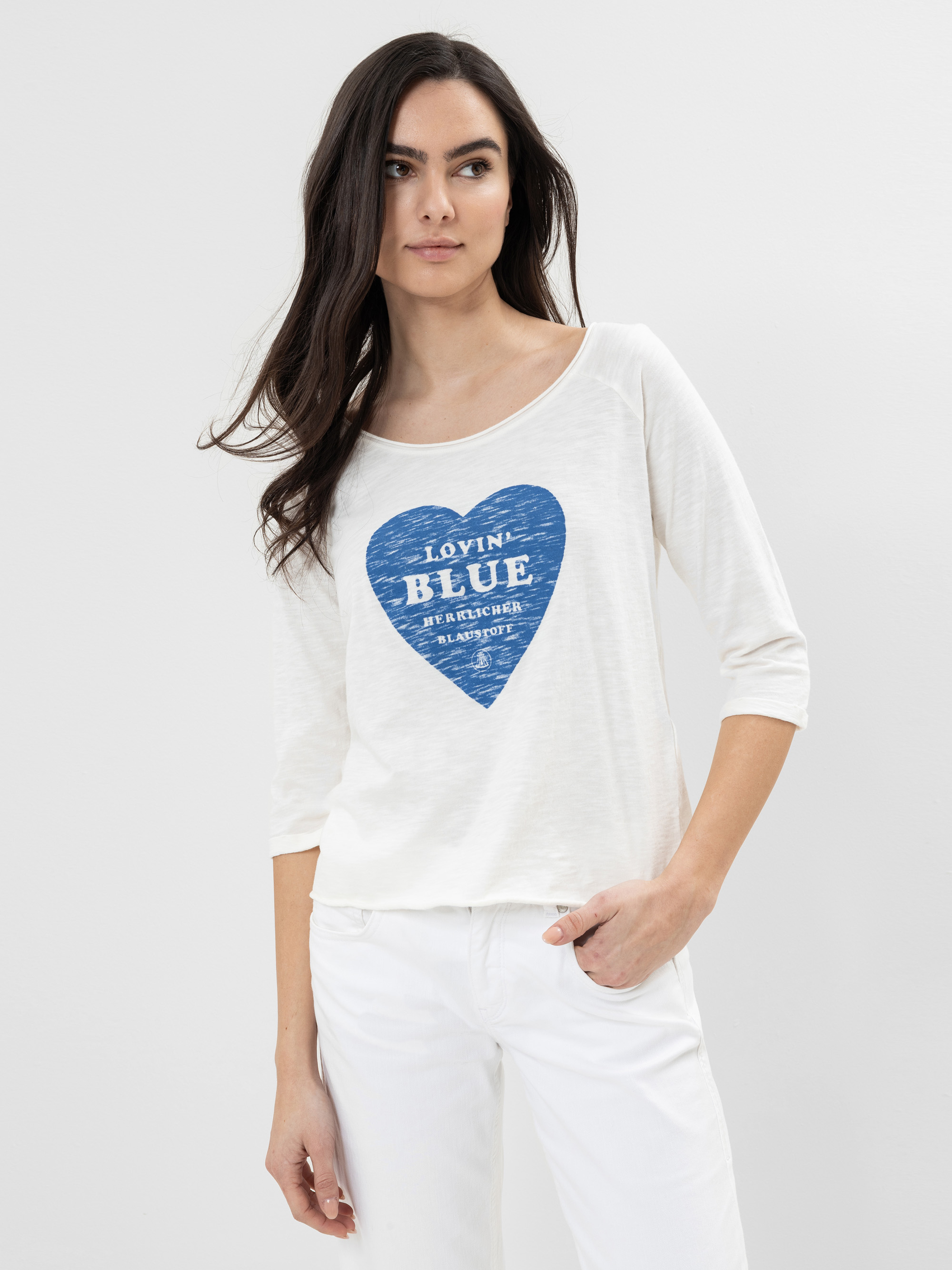 Eine Frau mit langen dunklen Haaren, die ein weißes Hemd mit einer blauen Herzgrafik und eine weiße Hose trägt, steht vor einem schlichten hellgrauen Hintergrund und schaut leicht zur Seite.