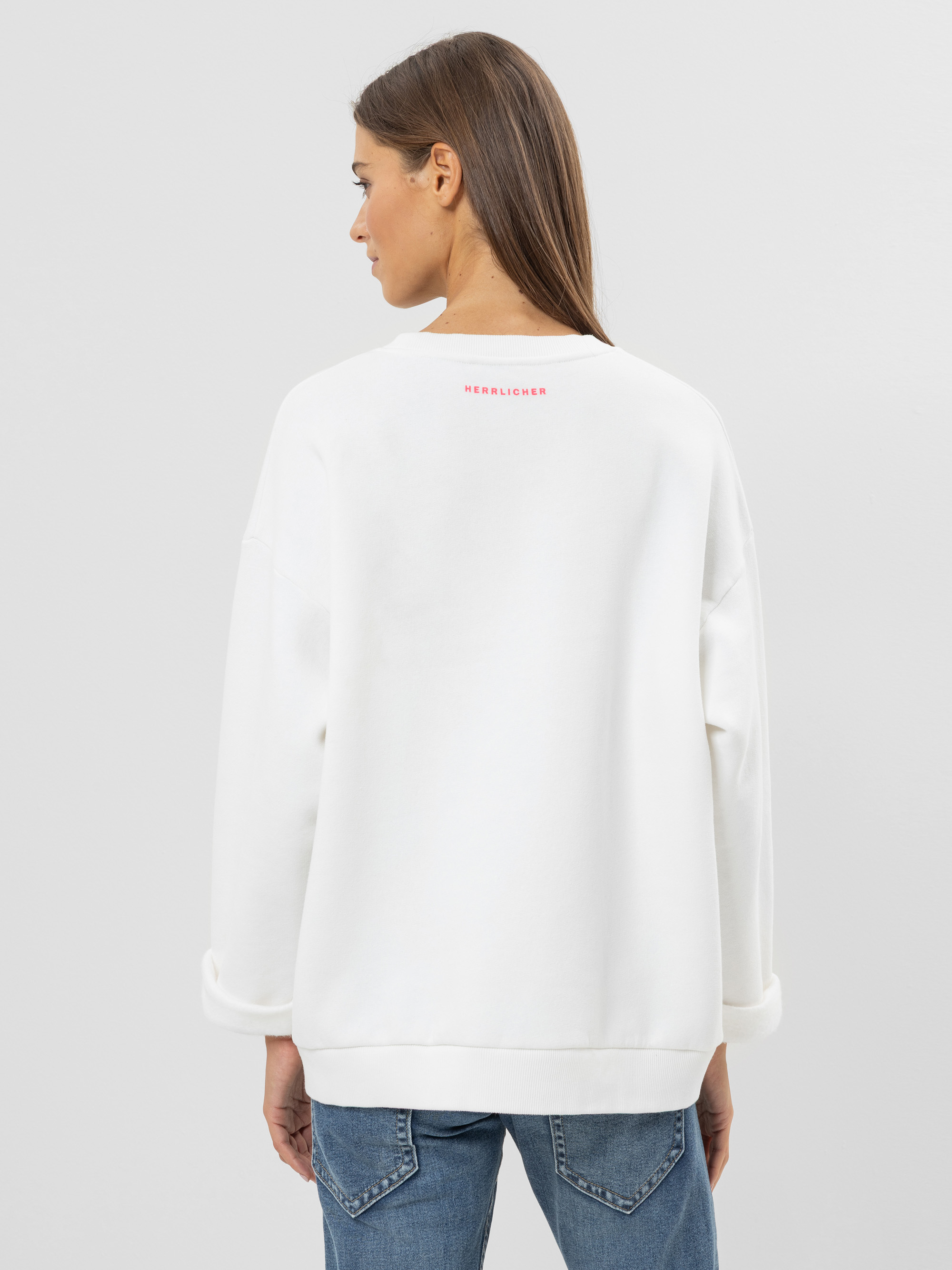 Eine Frau mit glattem braunem Haar ist von hinten zu sehen. Sie trägt ein lockeres weißes Sweatshirt mit aufgerollten Bündchen und blaue Jeans. Das Wort HERZLICHER ist in kleinen rosafarbenen Buchstaben in der Nähe des oberen Randes des Sweatshirts au