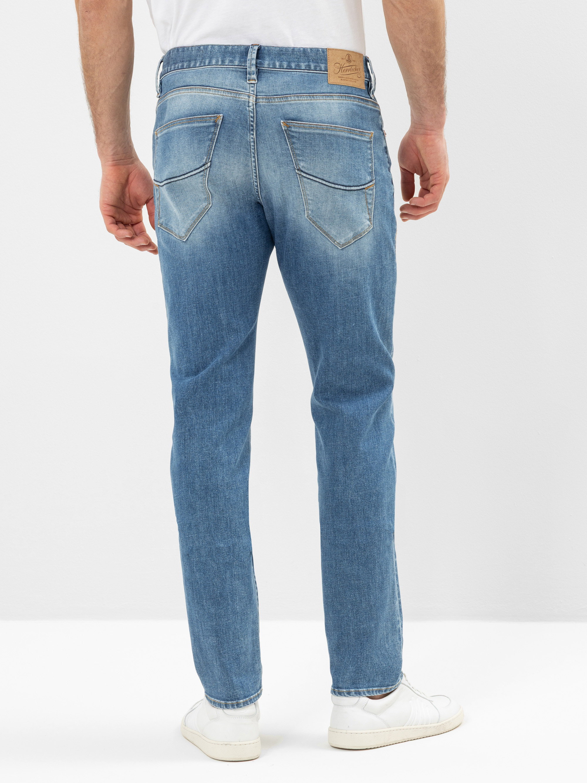 Ein Mann, der hellblaue Jeans und weiße Turnschuhe trägt, steht mit dem Rücken zur Kamera vor einem schlichten weißen Hintergrund. Die Jeans hat zwei Gesäßtaschen mit Nahtdetails und einen hellbraunen Aufnäher am Bund.