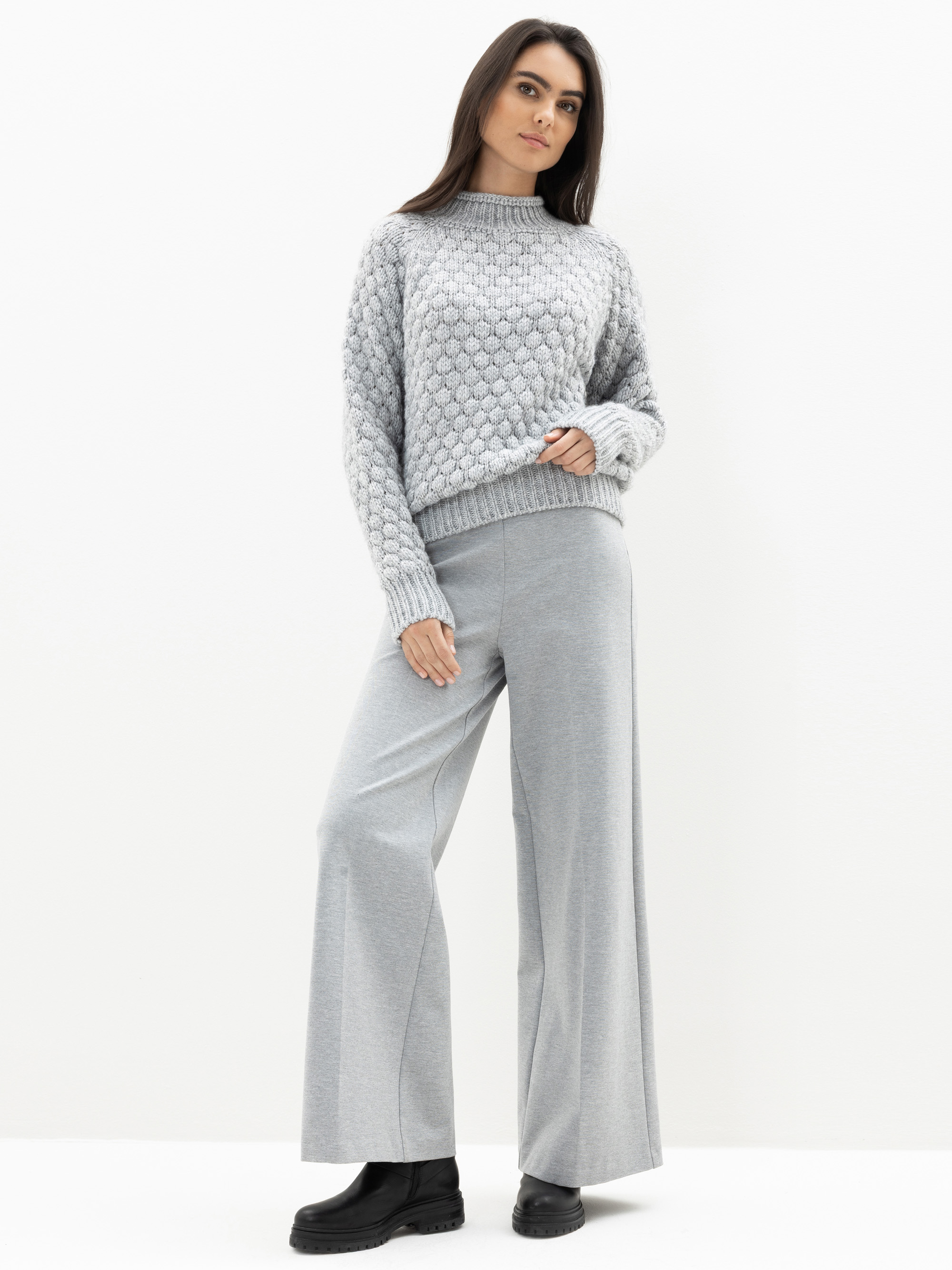 Eine Frau steht vor einem weißen Hintergrund und trägt einen hellgrauen strukturierten Pullover, eine graue Hose mit weitem Bein und schwarze Stiefel. Sie blickt leicht zur Seite, hat eine Hand in der Tasche und eine entspannte Körperhaltung.