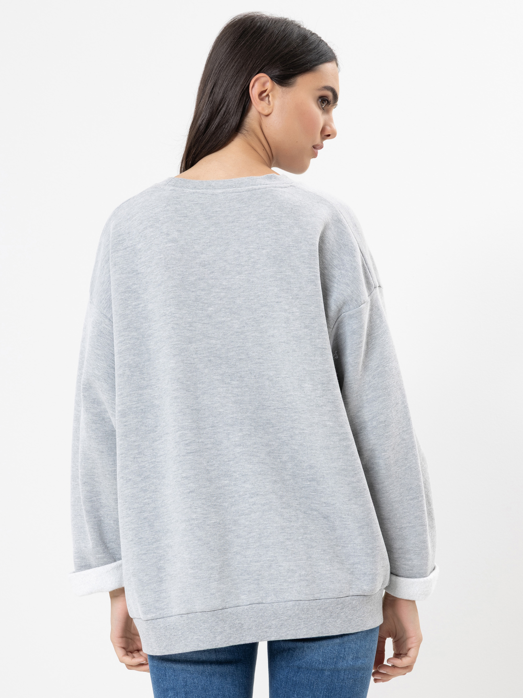 Eine Frau mit langen dunklen Haaren wird von hinten gezeigt. Sie trägt ein locker sitzendes hellgraues Sweatshirt und blaue Jeans vor einem schlichten weißen Hintergrund.