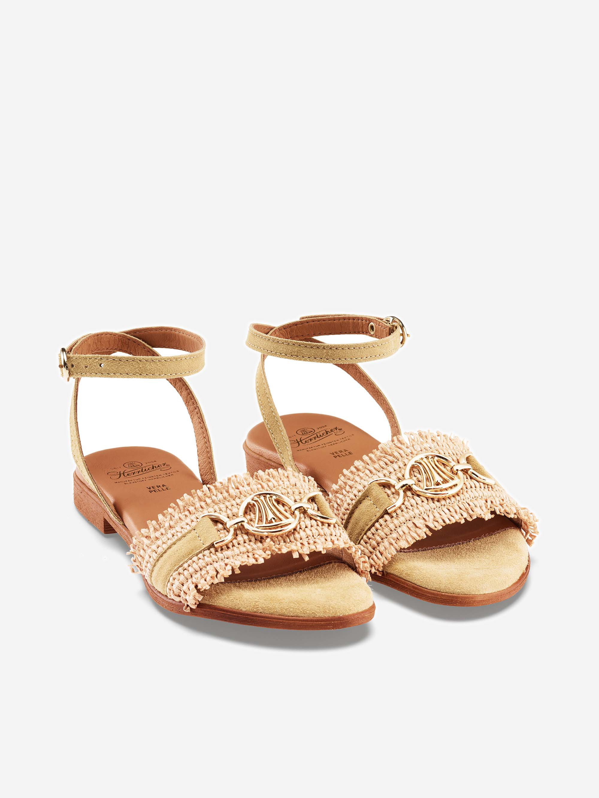 Ein Paar beigefarbene, zehenoffene Sandalen mit Knöchelriemen, goldenen Fransen und goldenen Zierbeschlägen an den vorderen Riemen, die vor einem schlichten weißen Hintergrund stehen.