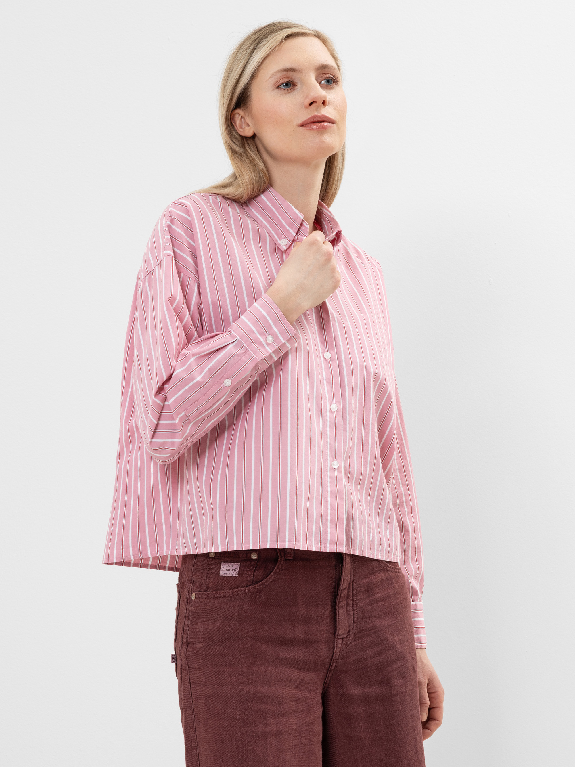 Eine Frau, die ein locker sitzendes, langärmeliges, rosa-weiß gestreiftes Hemd und eine kastanienbraune Hose trägt, steht vor einem schlichten weißen Hintergrund und blickt mit einem neutralen Ausdruck leicht nach oben.