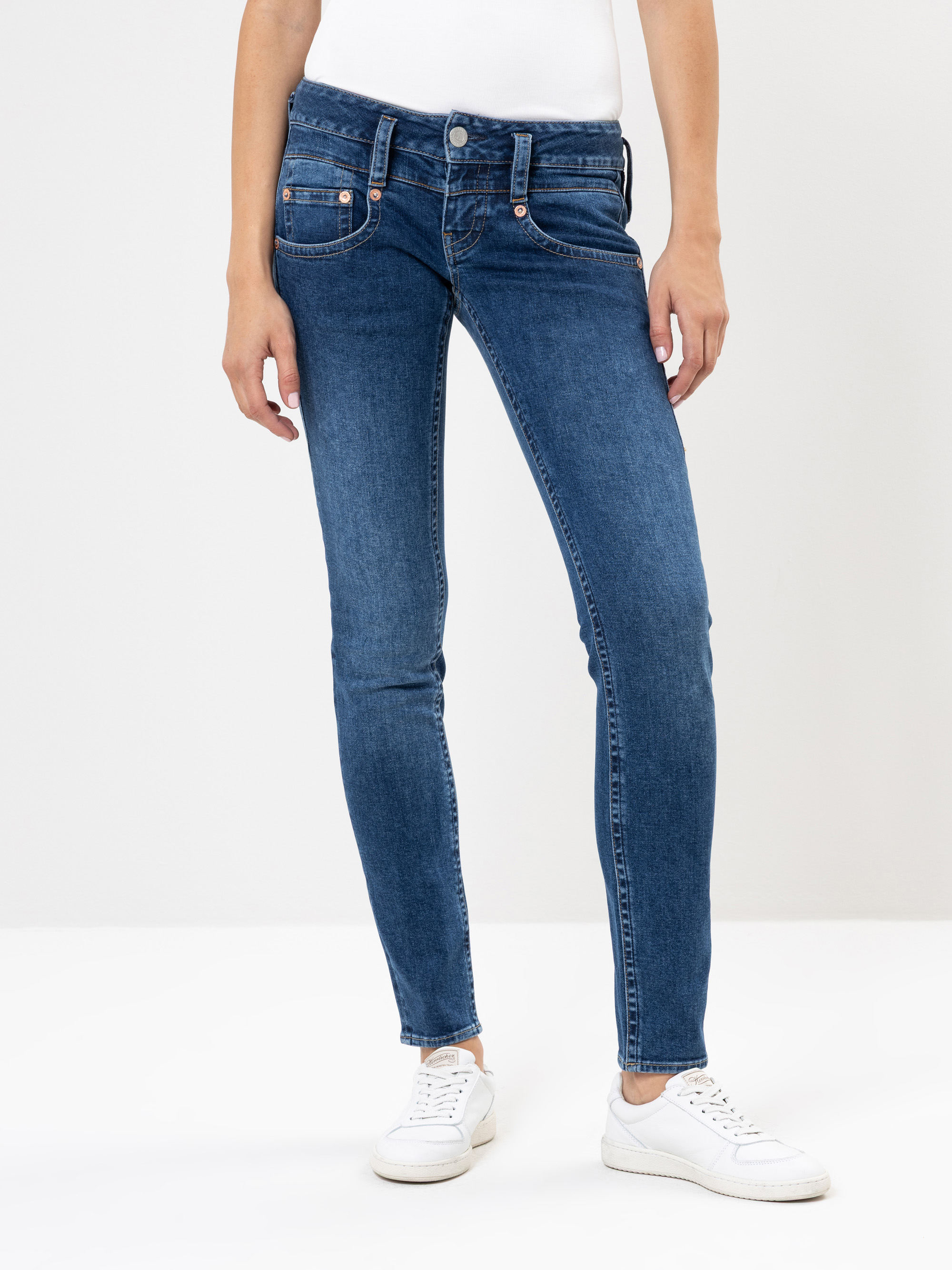 Eine Person in blauen Skinny-Jeans, einem weißen Oberteil und weißen Turnschuhen steht vor einem schlichten weißen Hintergrund. Das Bild konzentriert sich auf den Unterkörper und die Beine.