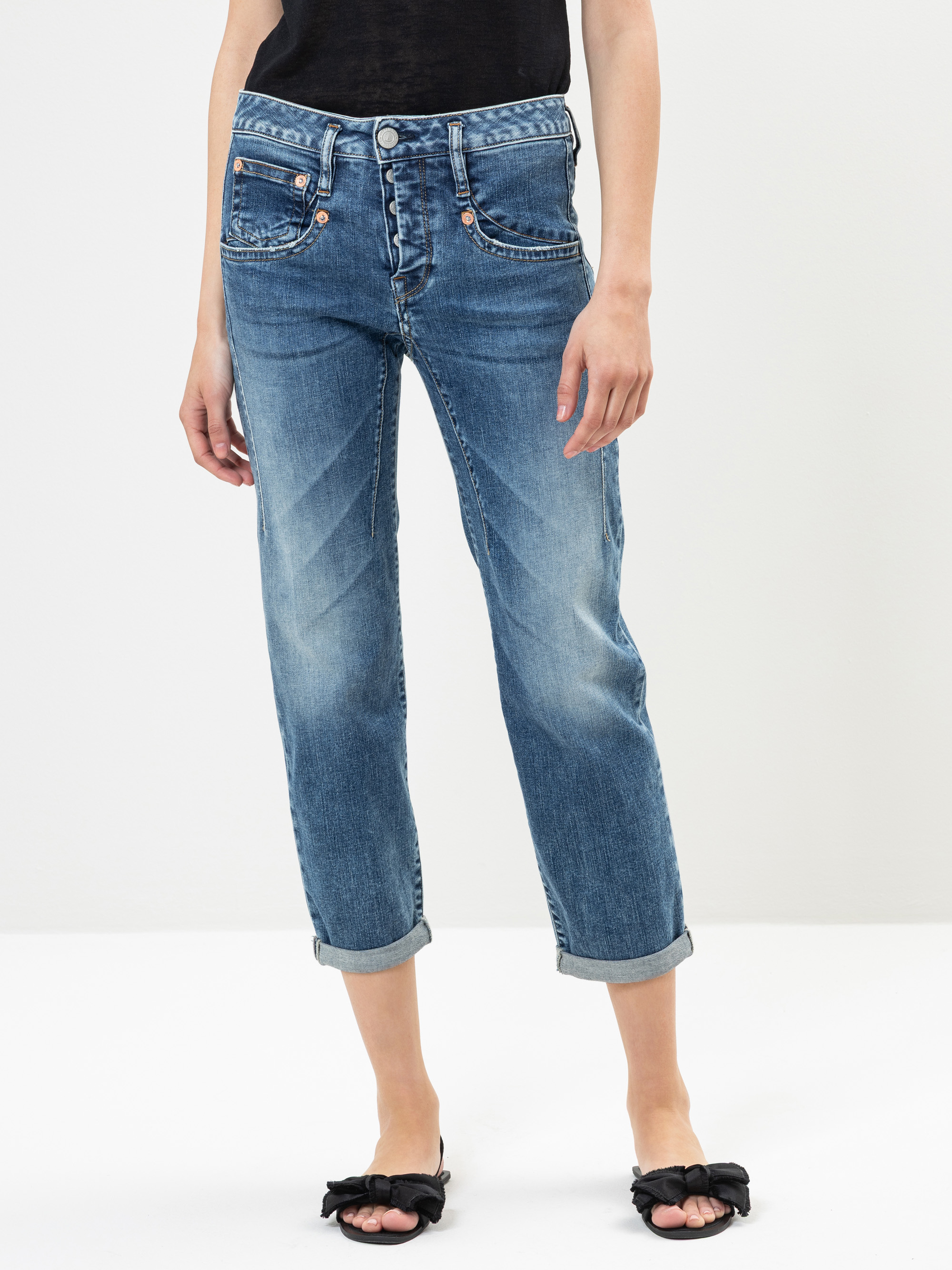 Eine Person, die blaue Jeans mit Bündchen und schwarze Sandalen mit Schleifen trägt, steht vor einem schlichten weißen Hintergrund. Ihre Hände liegen entspannt an der Seite, und ihr schwarzes Oberteil ist teilweise sichtbar.