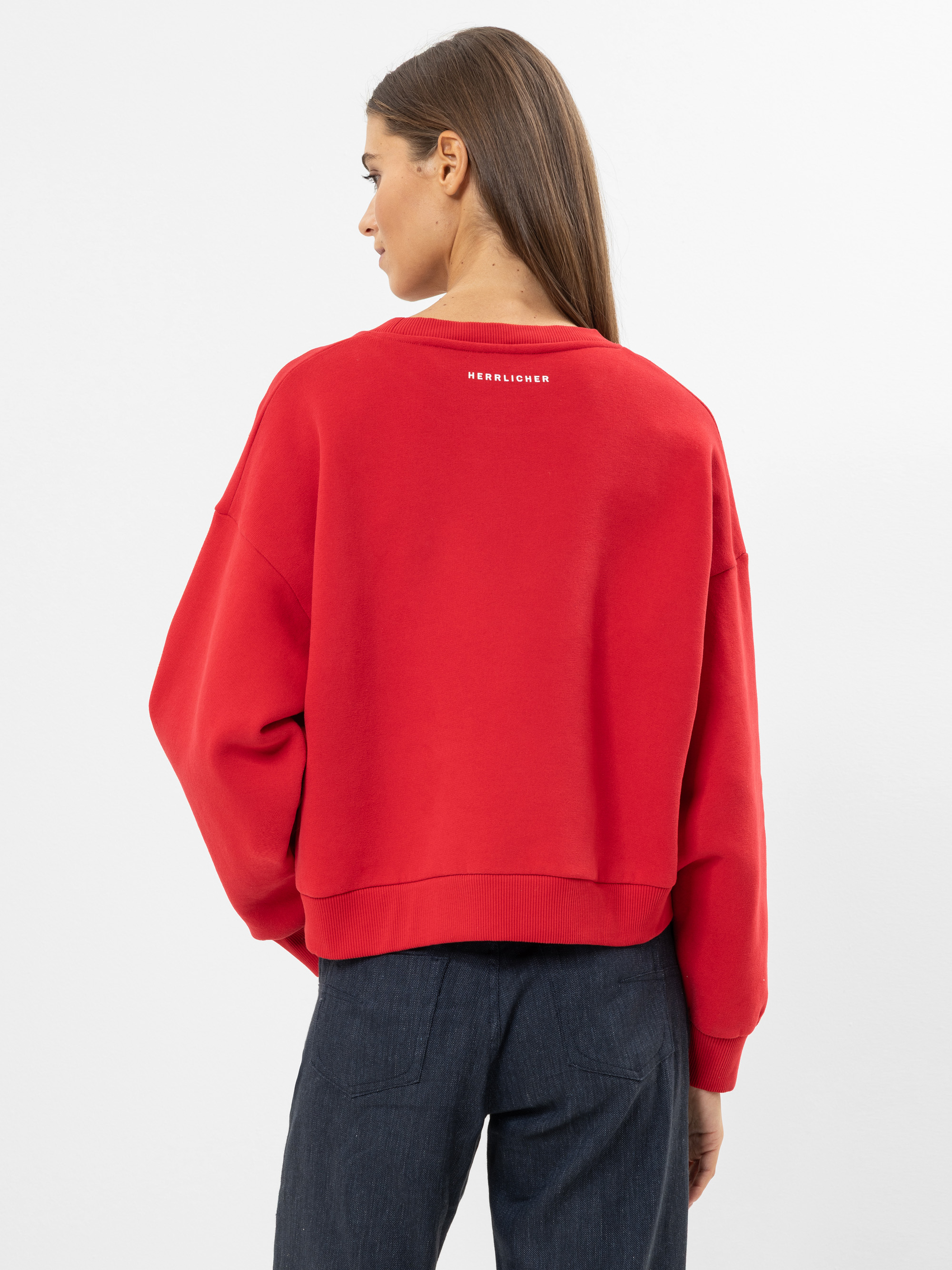 Eine Frau mit langen braunen Haaren wird von hinten gezeigt. Sie trägt ein locker sitzendes rotes Sweatshirt mit weißer Aufschrift auf dem oberen Rücken und eine dunkle Hose und steht vor einem schlichten hellen Hintergrund.