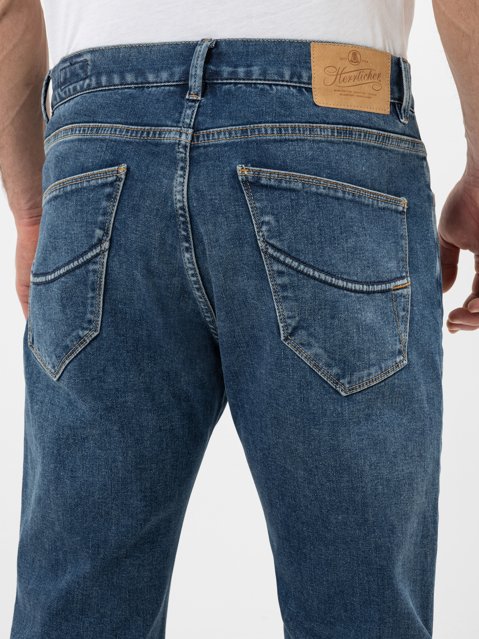 Nahaufnahme einer Person, die eine blaue Jeans trägt. Man sieht die Gesäßtaschen und einen braunen Markenaufnäher aus Leder am Hosenbund. Die Person trägt außerdem ein weißes Hemd, das in die Jeans gesteckt wird.