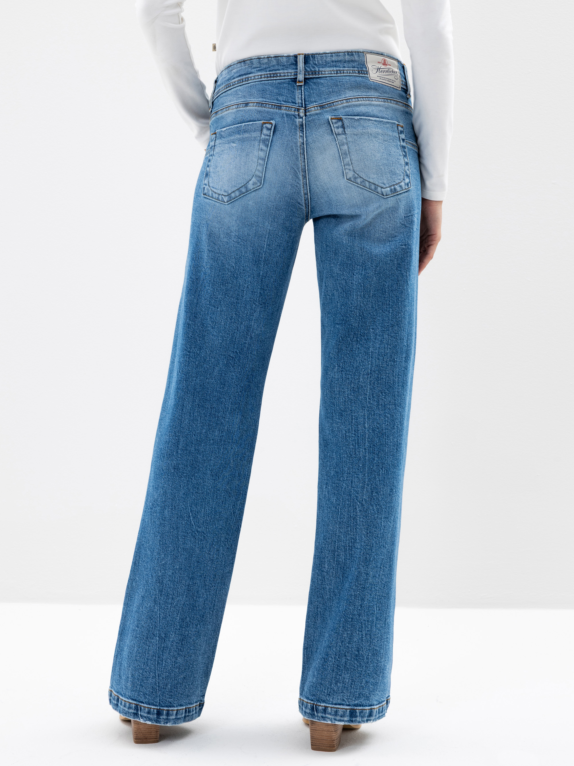 Eine Person, die eine hellblaue Jeans mit hoher Taille und weitem Bein und ein weißes langärmeliges Hemd trägt, steht mit dem Gesicht zur Seite und zeigt die Rückseite der Jeans mit zwei Gesäßtaschen und braunen Schuhen mit Absatz.