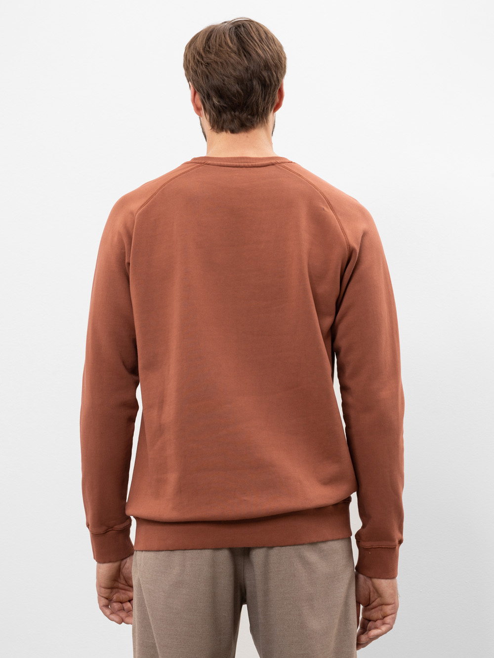 Ein Mann mit kurzen braunen Haaren steht mit dem Rücken zur Kamera, trägt ein rostfarbenes Sweatshirt und eine hellbraune Hose vor einem schlichten weißen Hintergrund.