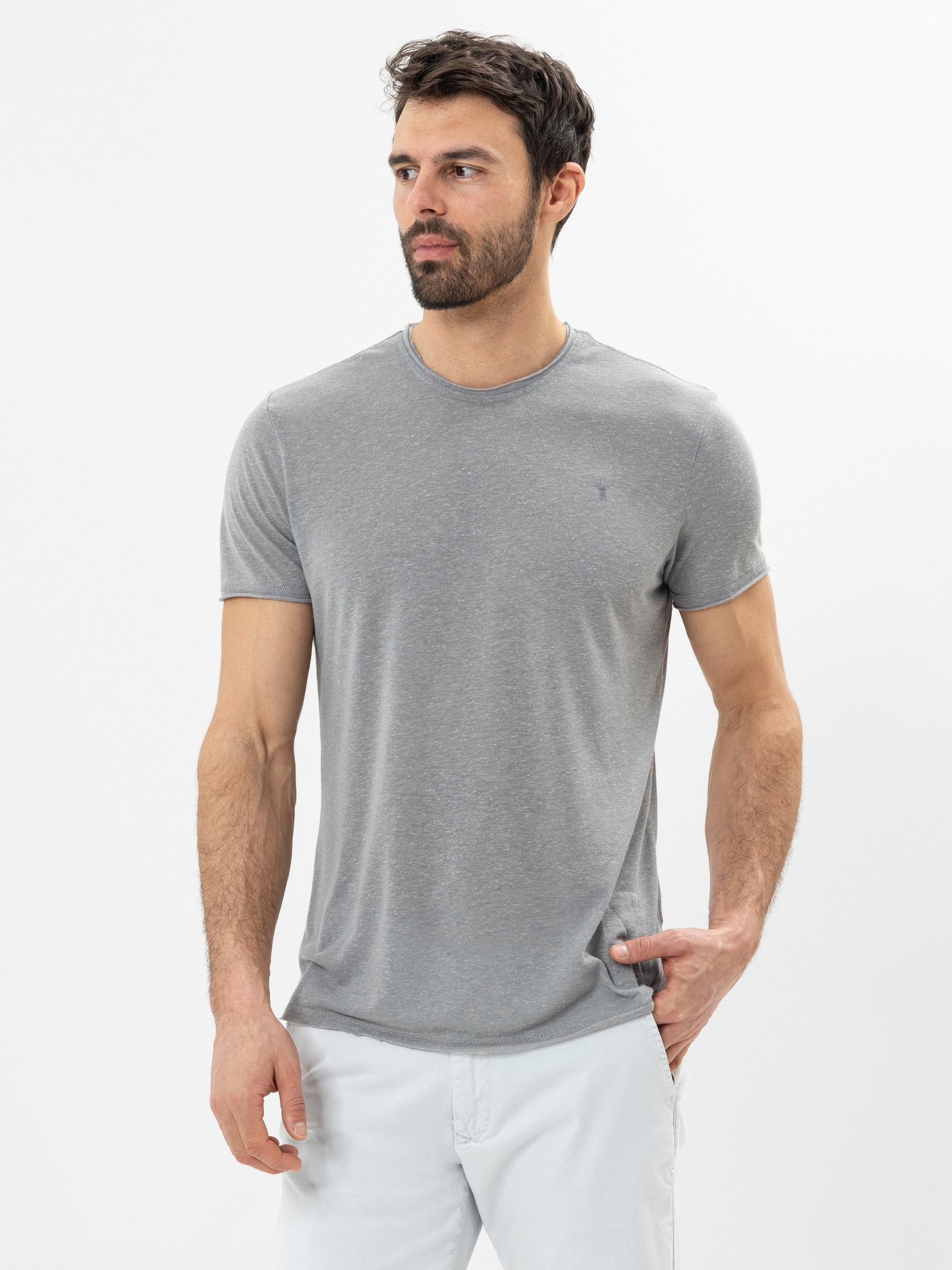 Ein Mann mit kurzen dunklen Haaren und einem Bart trägt ein graues T-Shirt und eine weiße Hose. Er steht vor einem schlichten weißen Hintergrund und blickt leicht nach links.
