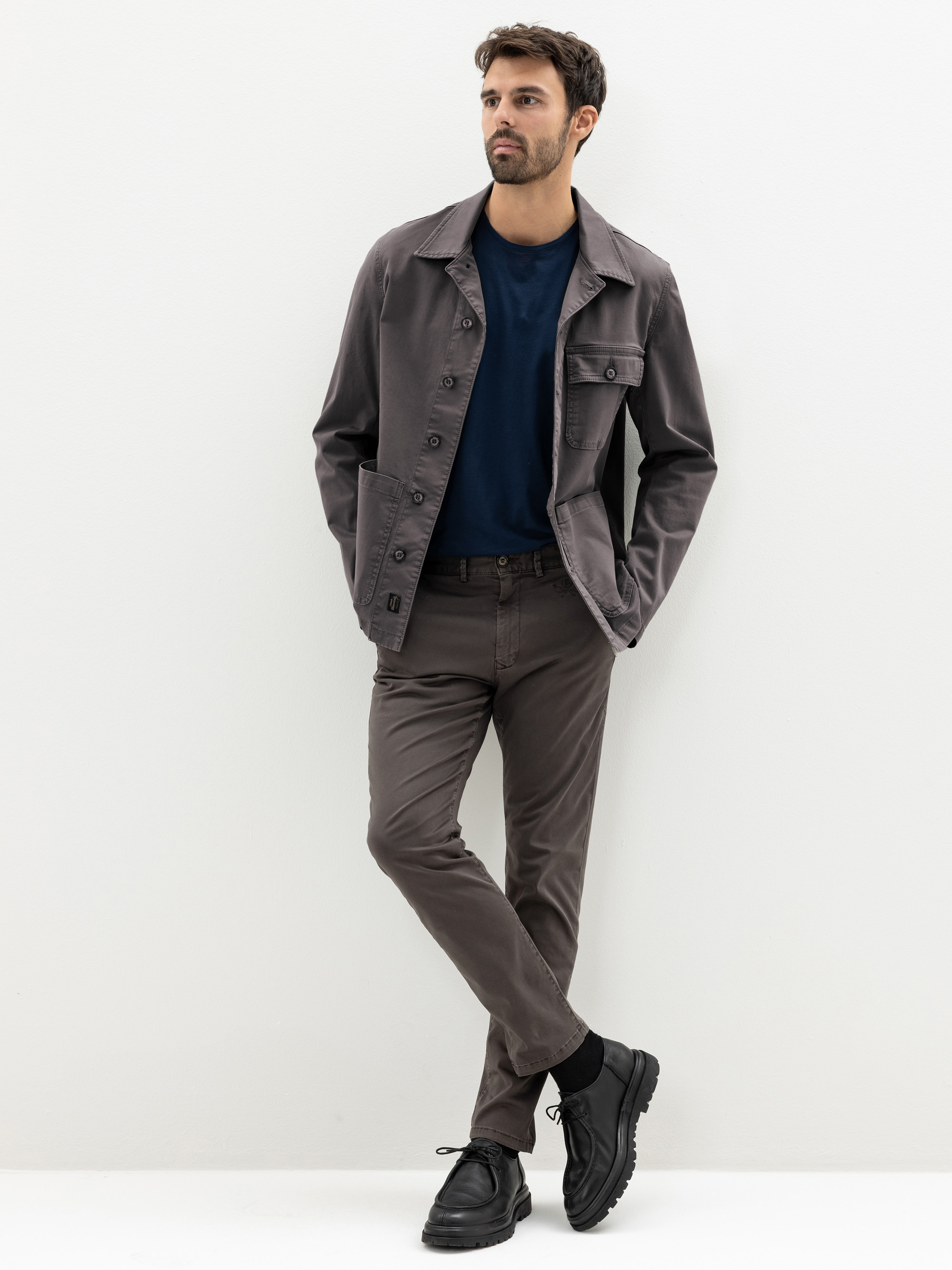 Ein Mann steht vor einem weißen Hintergrund und trägt eine graue Jacke, ein dunkelblaues T-Shirt, eine graue Hose und schwarze Schuhe. Er hat kurze Haare und einen Bart, eine Hand steckt in der Tasche und ein Bein ist über das andere geschlagen.