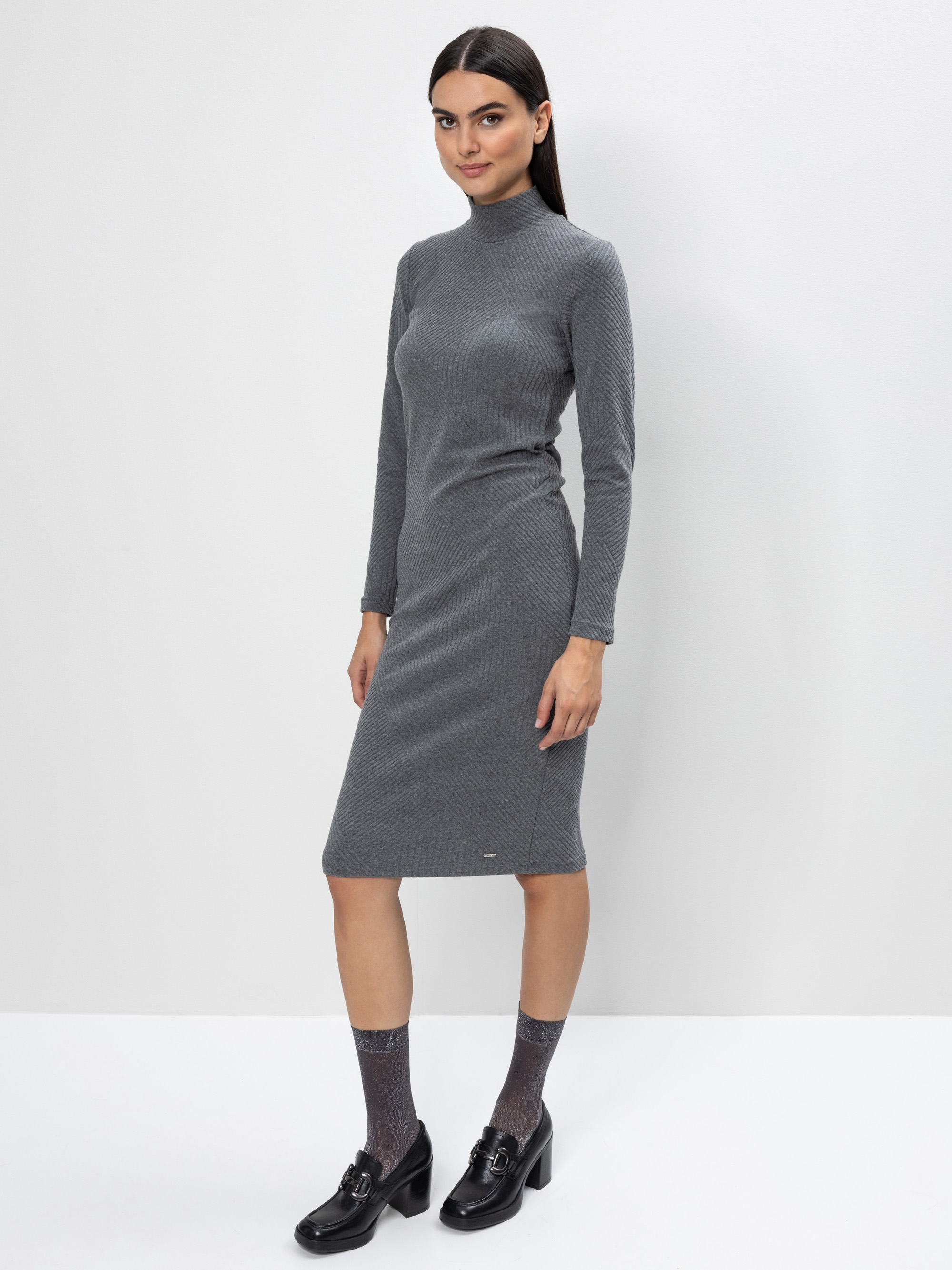 Eine Frau mit langen dunklen Haaren, die ein graues, geripptes Rollkragenkleid mit langen Ärmeln, graue Socken und schwarze Halbschuhe mit Absatz trägt, steht vor einem schlichten weißen Hintergrund.