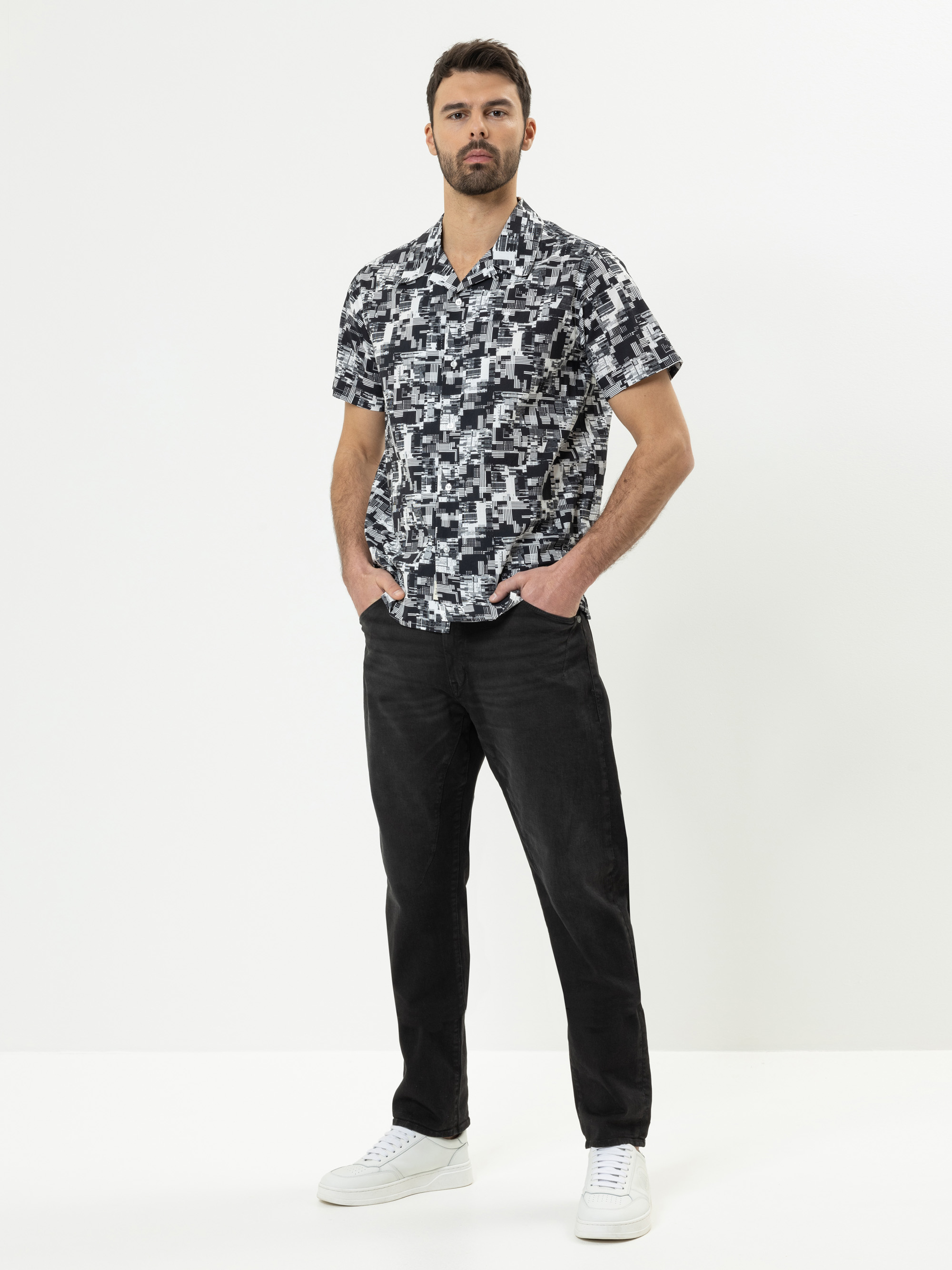 Ein Mann steht vor einem weißen Hintergrund und trägt ein schwarz-weißes, geometrisch gemustertes, kurzärmeliges Hemd, eine schwarze Hose und weiße Turnschuhe. Er hat dunkles Haar, einen Bart und die Hände in den Taschen.