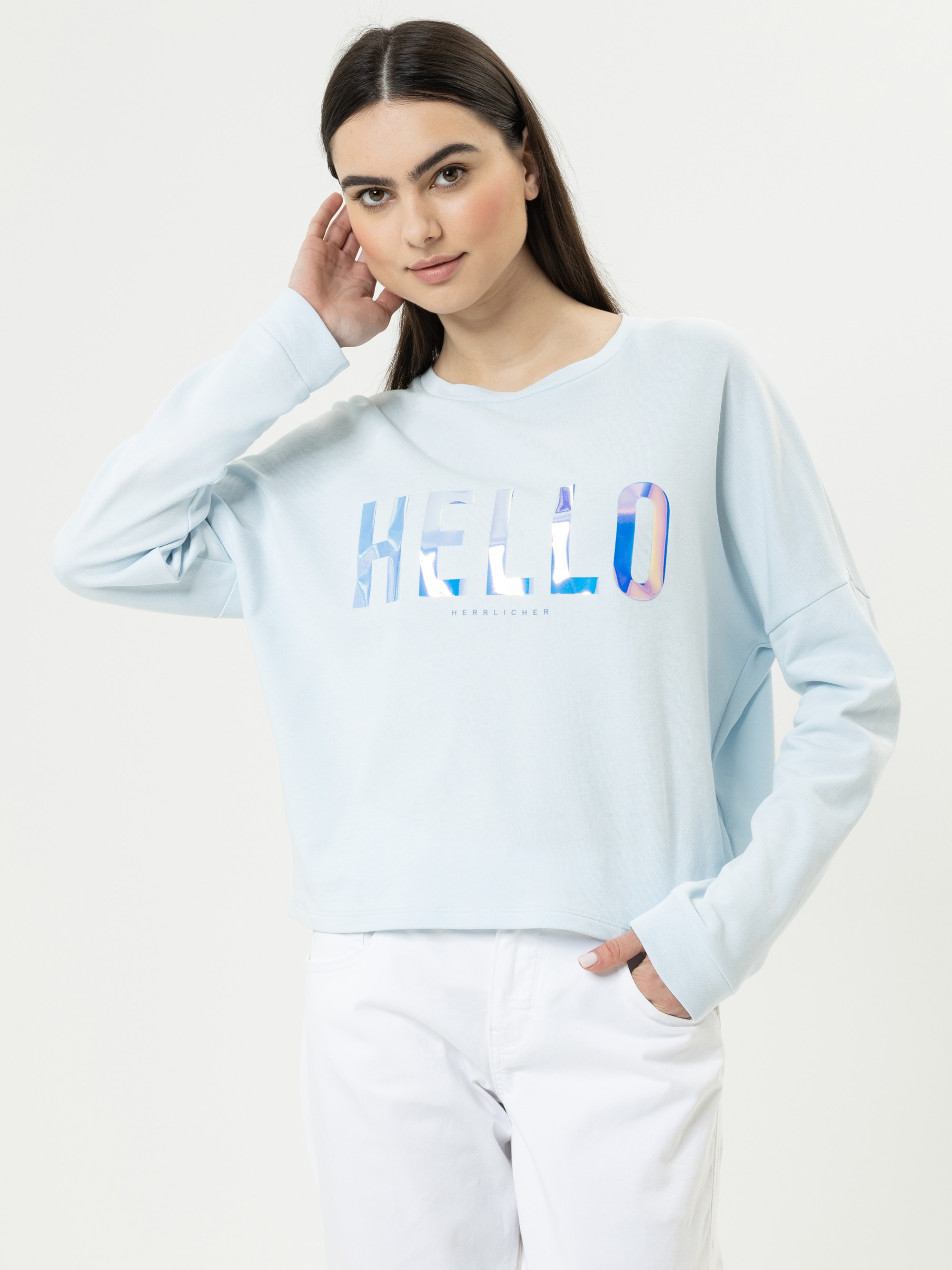 Eine Frau mit langen dunklen Haaren trägt ein hellblaues Sweatshirt, auf dem in glänzenden Buchstaben HELLO steht, und eine weiße Hose. Sie steht vor einem schlichten hellen Hintergrund, eine Hand am Ohr, die andere in der Tasche.