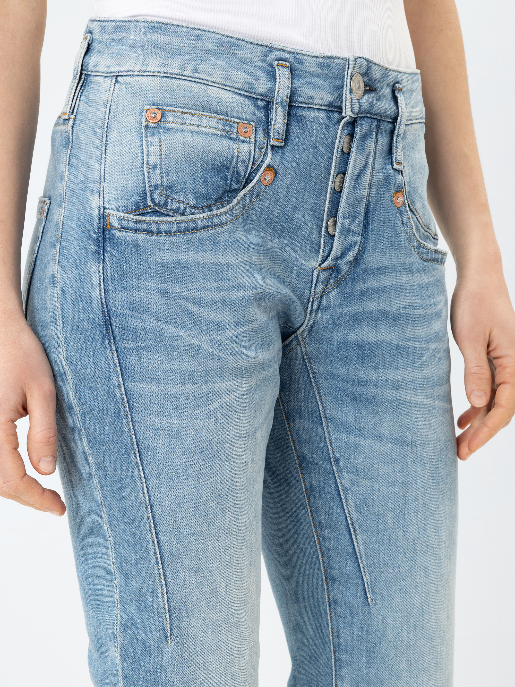 Eine Person trägt eine hellblaue Jeans mit hoher Taille und sichtbaren Nähten auf der Vorderseite und freiliegenden Knöpfen, dazu ein weißes Oberteil zum Hineinstecken. Nur die Taille und die Oberschenkel sind sichtbar.
