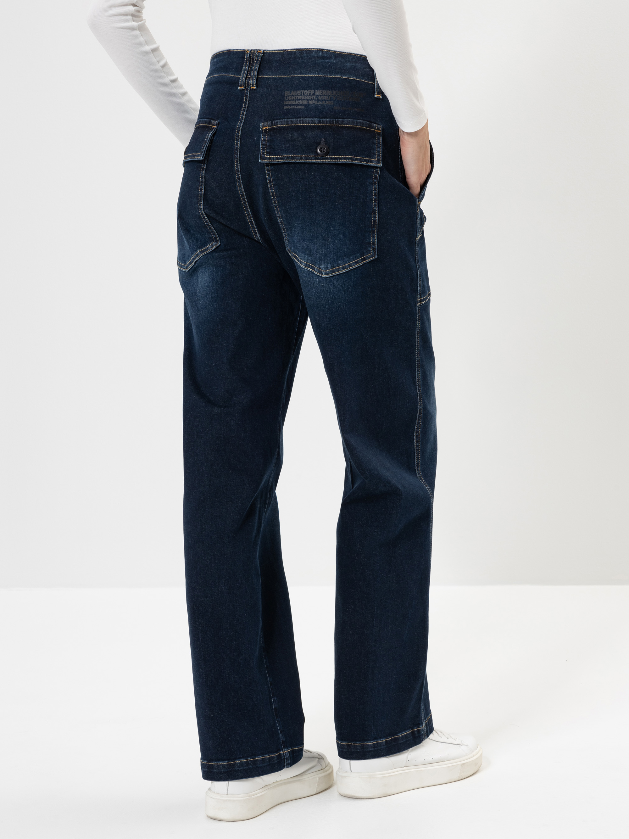 Eine Person, die dunkelblaue Jeans mit großen Gesäßtaschen und weiße Turnschuhe trägt, steht mit einer Hand in der Hosentasche, von der Kamera abgewandt, vor einem schlichten weißen Hintergrund.