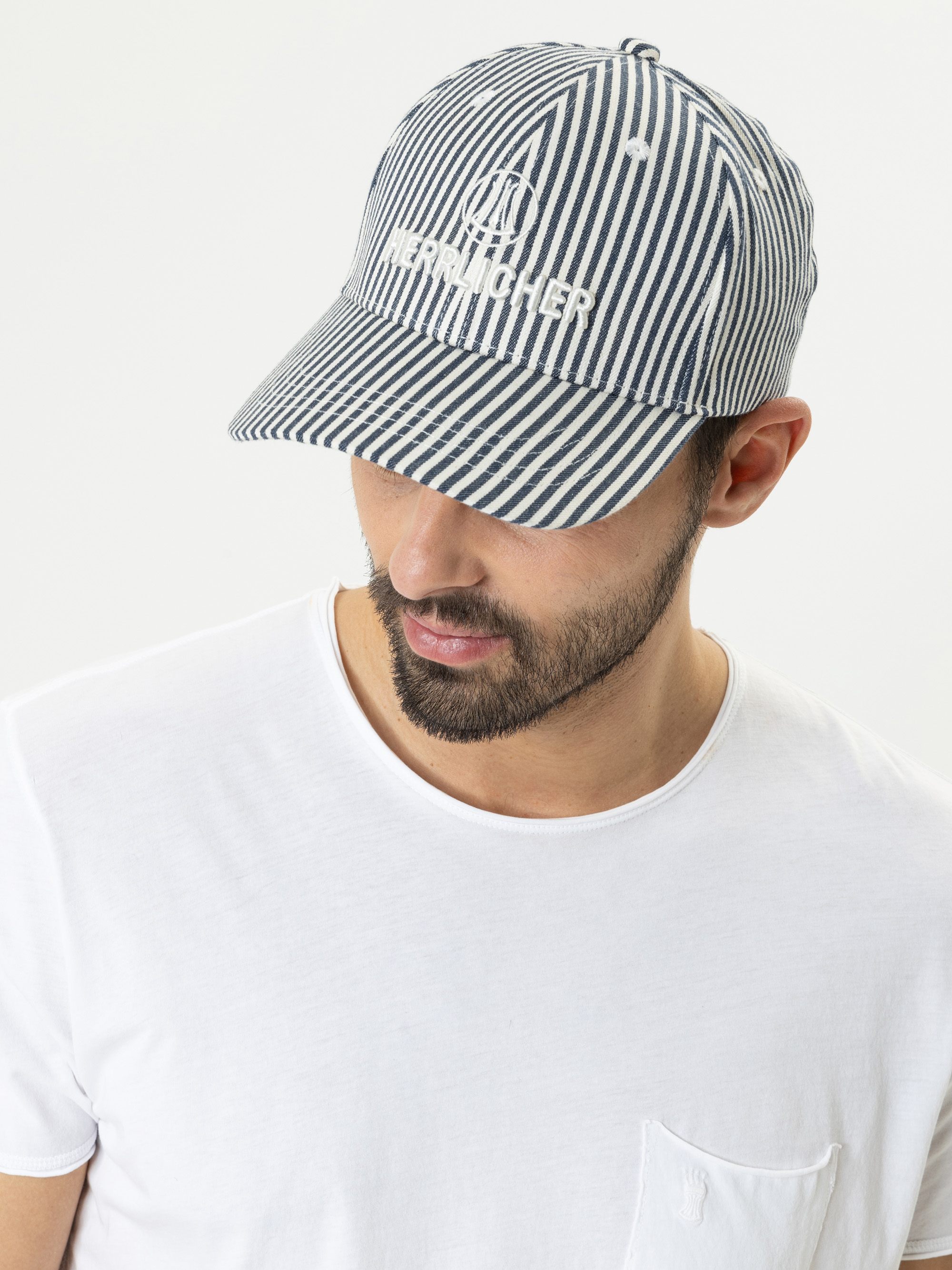 Ein Mann, der ein weißes T-Shirt und eine blau-weiß gestreifte Baseballmütze mit der Aufschrift HERHICHER auf der Vorderseite trägt, blickt vor einem schlichten weißen Hintergrund nach unten.