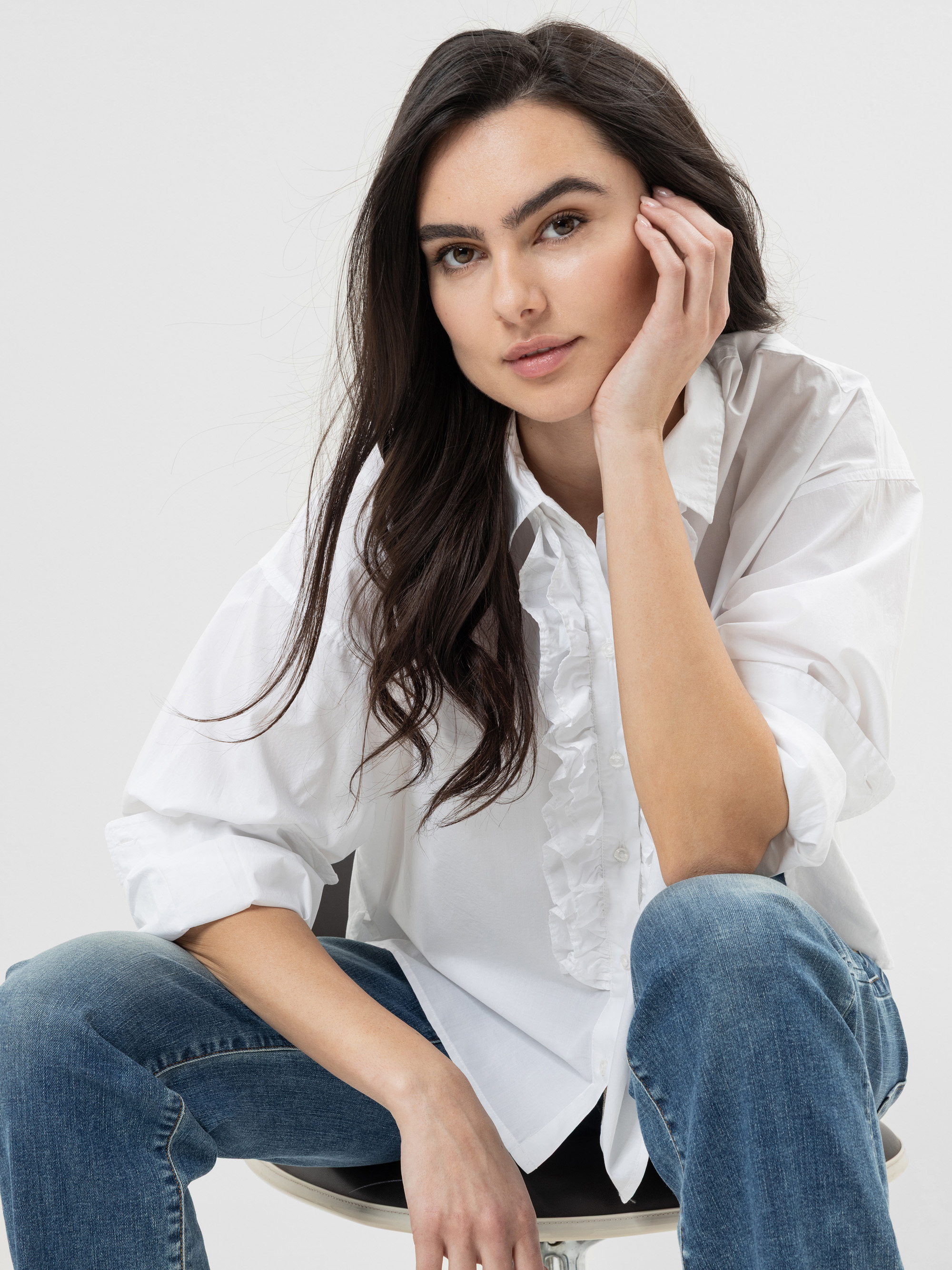 Eine Frau mit langen dunklen Haaren, die ein weißes Rüschenhemd und eine blaue Jeans trägt, sitzt auf einem Stuhl und stützt ihr Gesicht auf ihre Hand, während sie vor einem schlichten hellen Hintergrund selbstbewusst in die Kamera schaut.