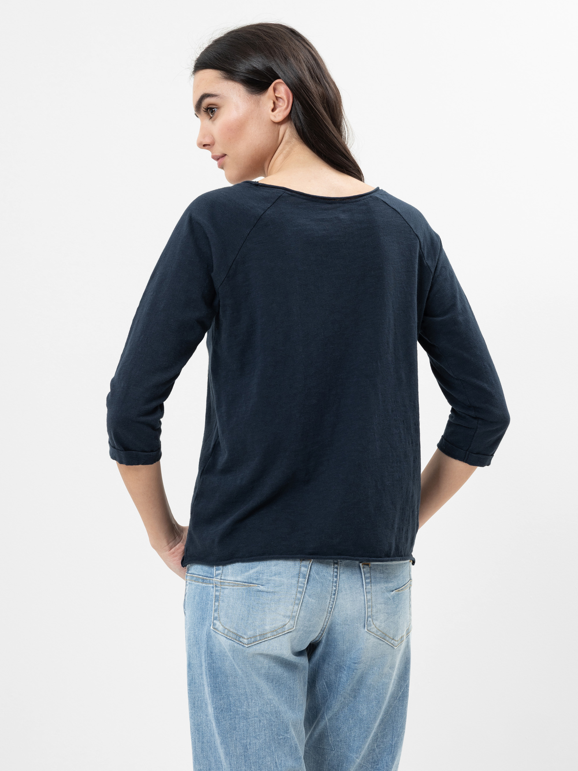 Eine Frau mit langen dunklen Haaren steht mit dem Gesicht nach hinten, trägt ein marineblaues langärmeliges Oberteil und hellblaue Jeans vor einem schlichten weißen Hintergrund.