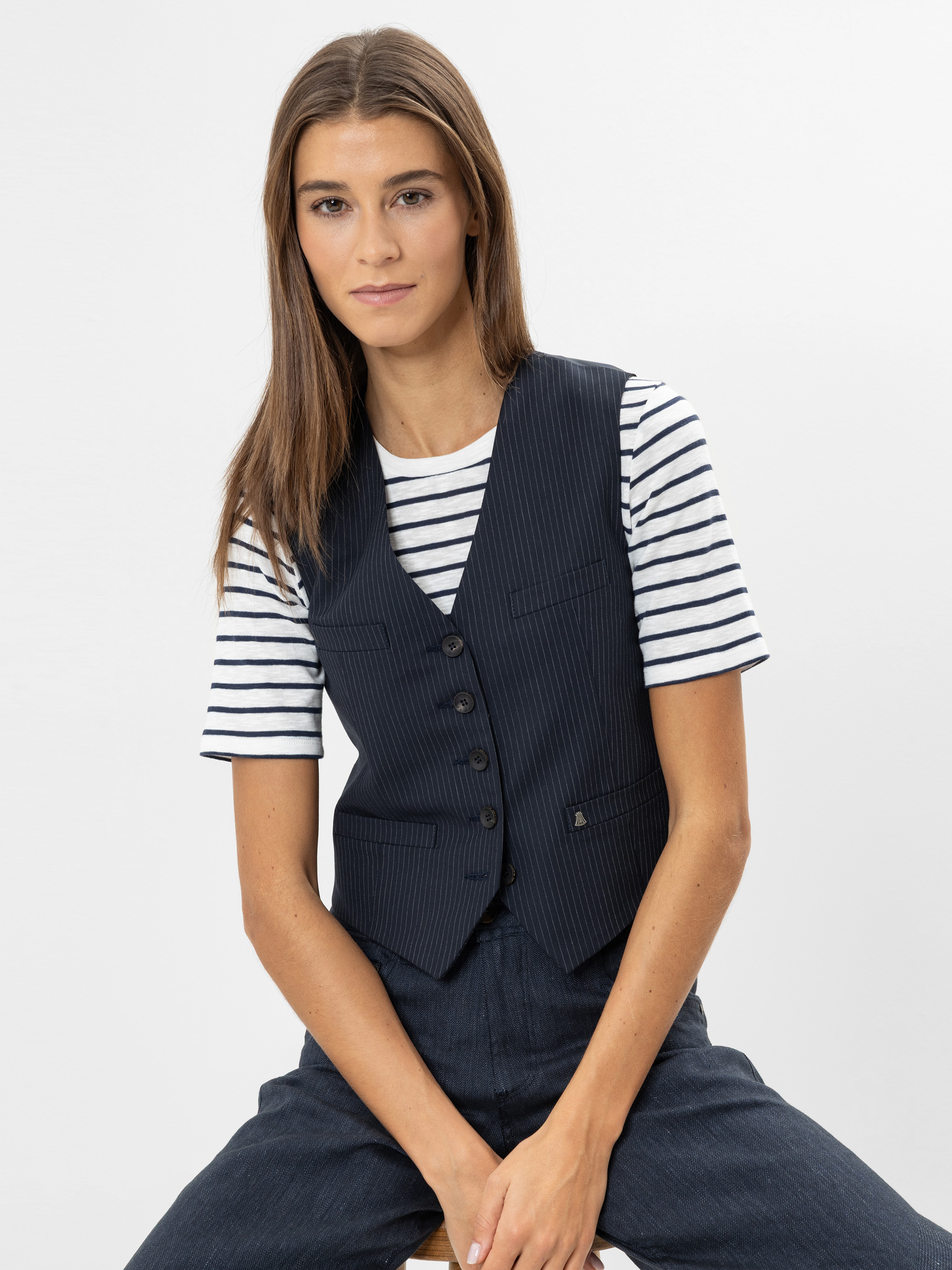 Eine Person mit langen braunen Haaren sitzt auf einem Hocker und trägt eine marineblaue Nadelstreifenweste über einem weiß und marineblau gestreiften T-Shirt und eine passende marineblaue Hose vor einem schlichten weißen Hintergrund.
