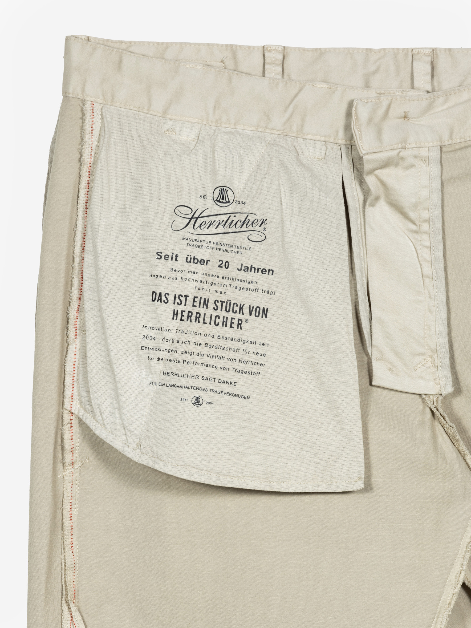 Nahaufnahme der Innenseite einer beigen Hose, die eine große weiße Tasche mit schwarzem Text in deutscher Sprache zeigt, darunter der Markenname Herrlicher und Informationen über die Qualität und Einzigartigkeit des Kleidungsstücks.