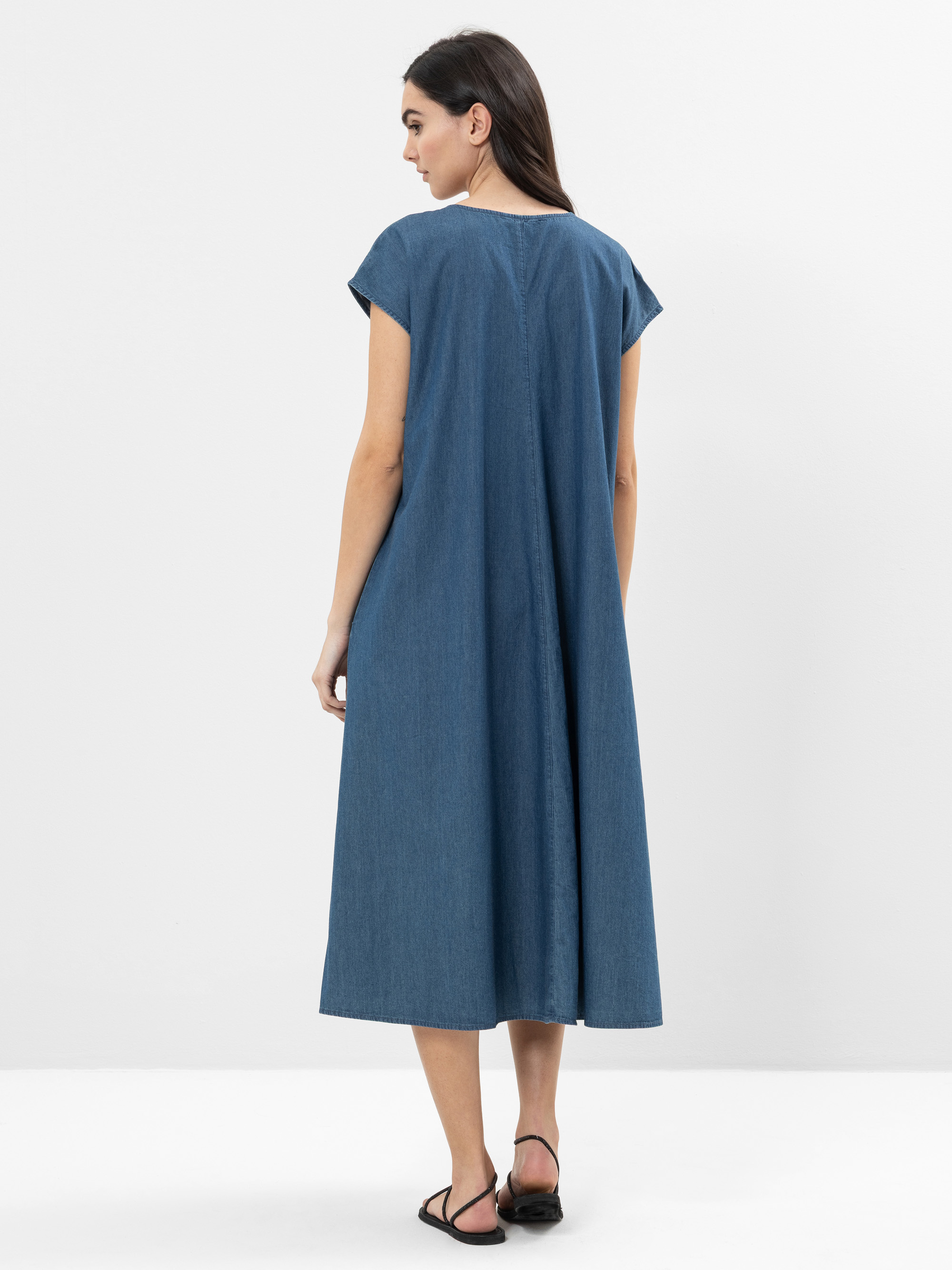 Stehende und abgewandte Frau, die ein locker sitzendes, kurzärmeliges, knöchellanges blaues Jeanskleid und schwarze Sandalen vor einem schlichten weißen Hintergrund trägt.