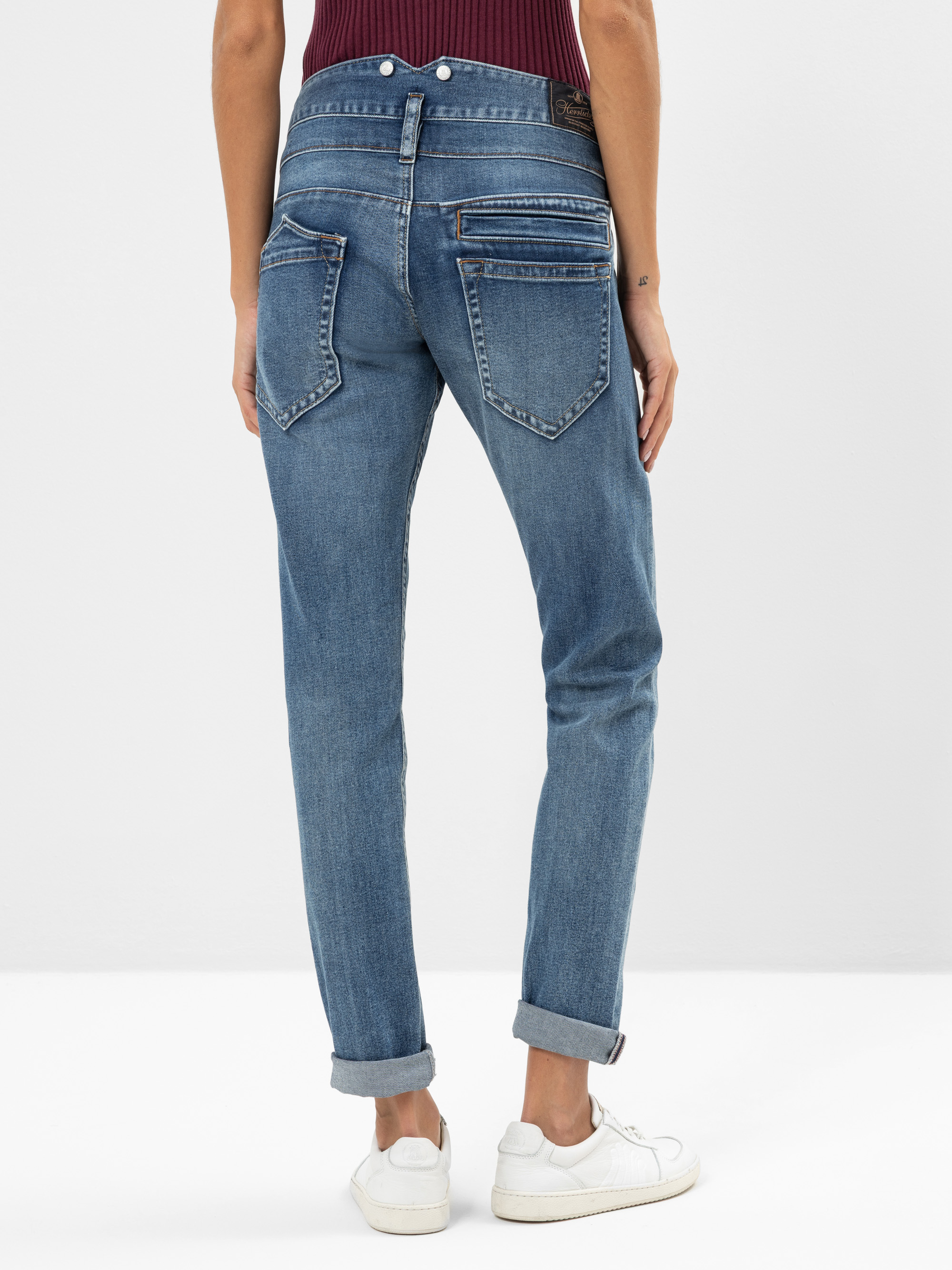 Die Person trägt eine blaue Jeans mit gekrempelten Bündchen und weiße Turnschuhe, von hinten gesehen. Die Jeans hat einzigartige diagonale Nähte und Details mit Reißverschluss an den Gesäßtaschen. Die Person steht vor einem schlichten, hellen Hinte