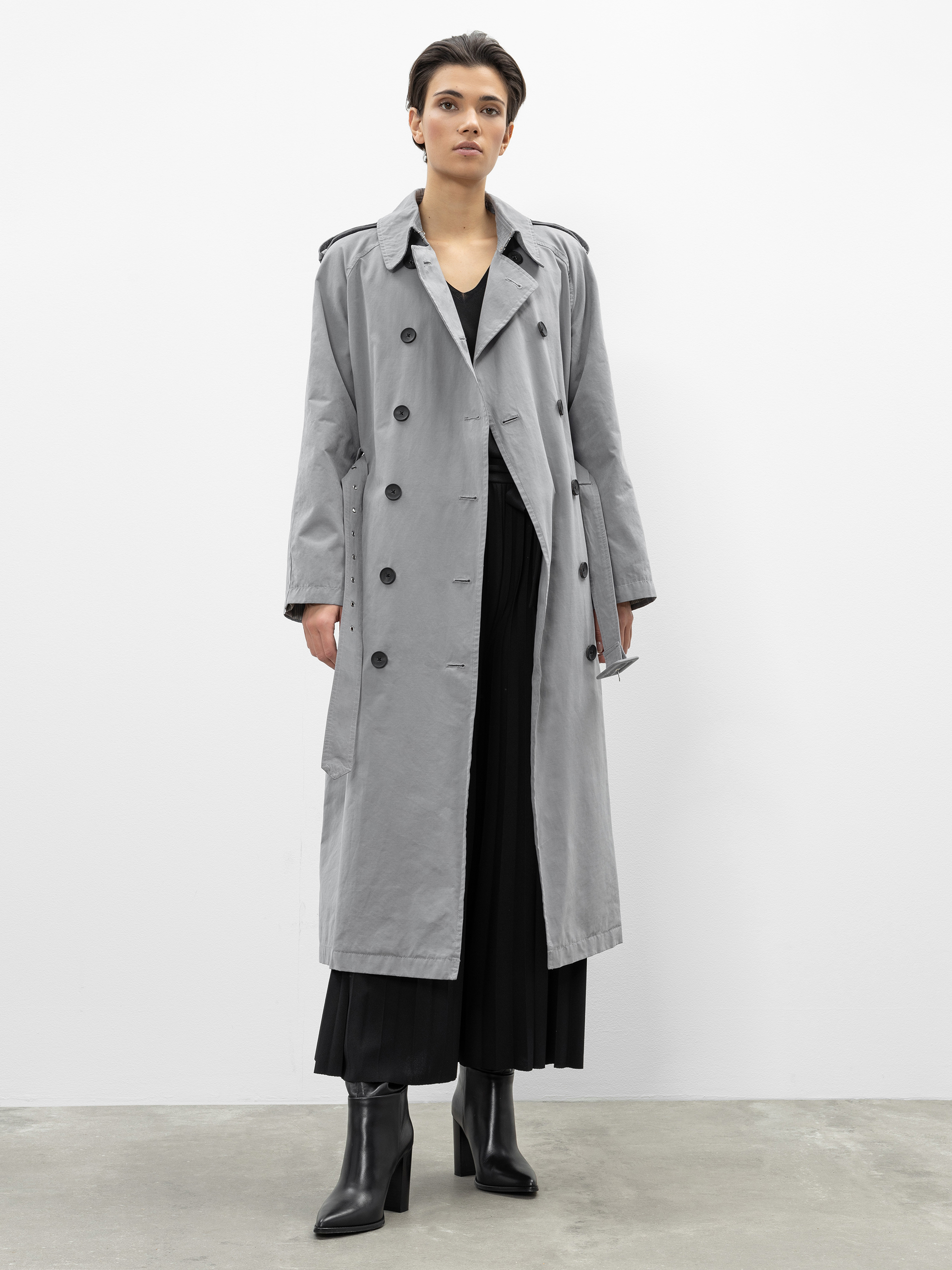 Eine Person mit kurzen dunklen Haaren trägt einen langen grauen Trenchcoat über einem schwarzen Outfit und schwarze hochhackige Stiefel und steht vor einer schlichten weißen Wand auf einem Betonboden.