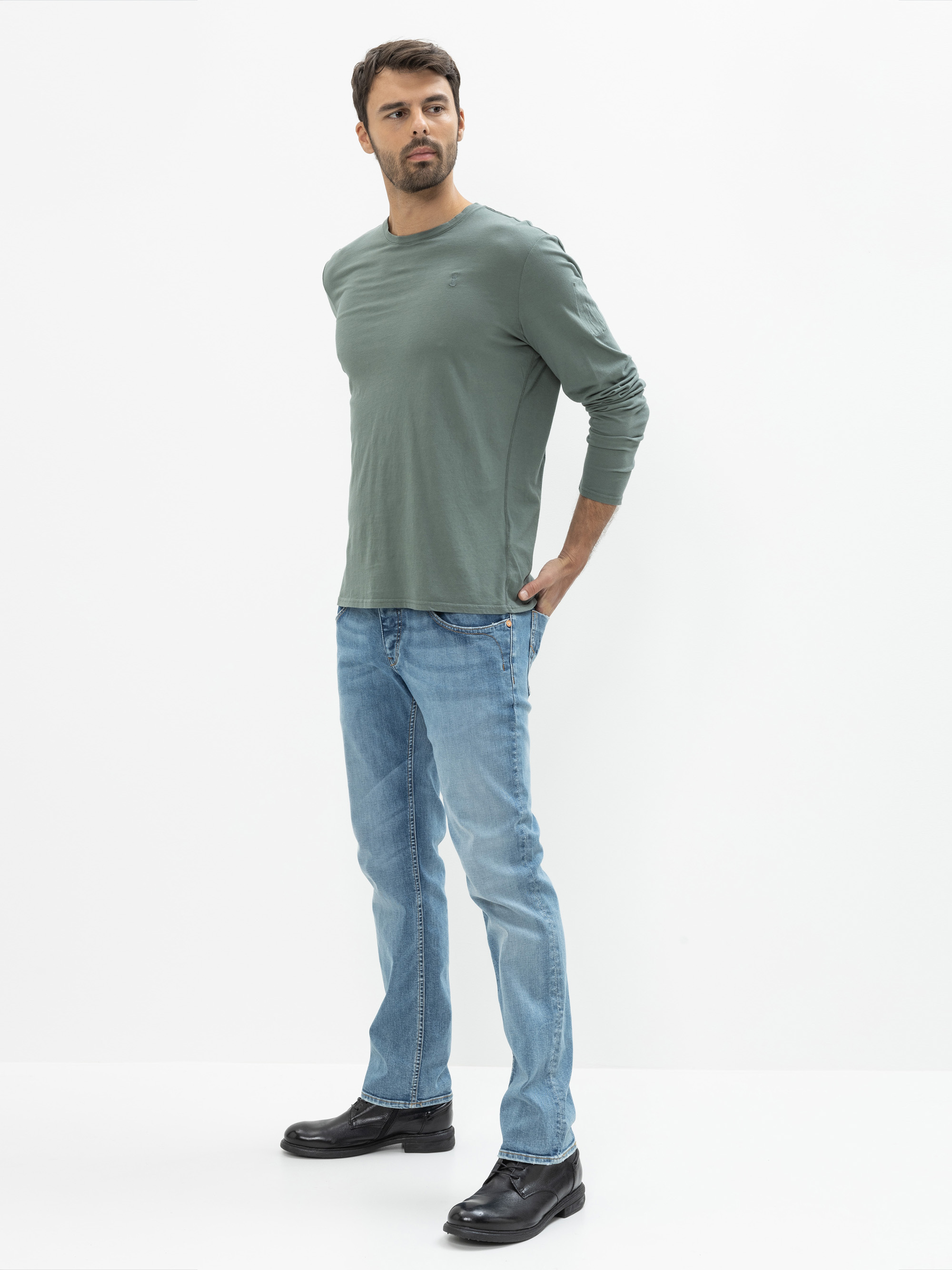 Ein Mann mit kurzen dunklen Haaren und einem Bart trägt ein salbeigrünes langärmeliges Hemd, hellblaue Jeans und schwarze Schuhe. Er steht vor einem schlichten weißen Hintergrund und blickt leicht zur Seite, eine Hand in die Hüfte gestützt.