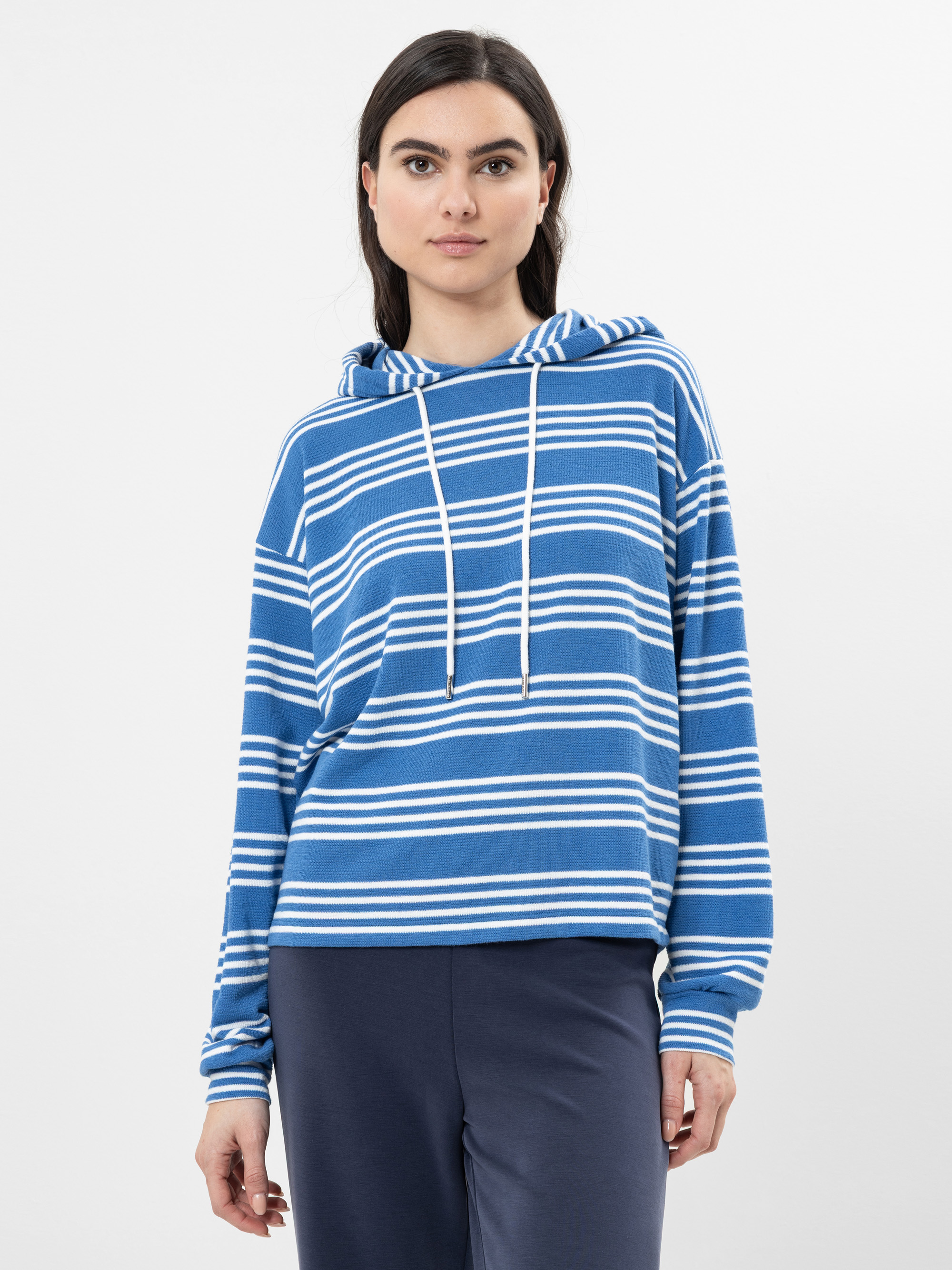 Eine Frau mit langen dunklen Haaren, die einen blau-weiß gestreiften Kapuzenpullover mit Kordelzug und eine marineblaue Hose trägt, steht vor einem schlichten weißen Hintergrund.