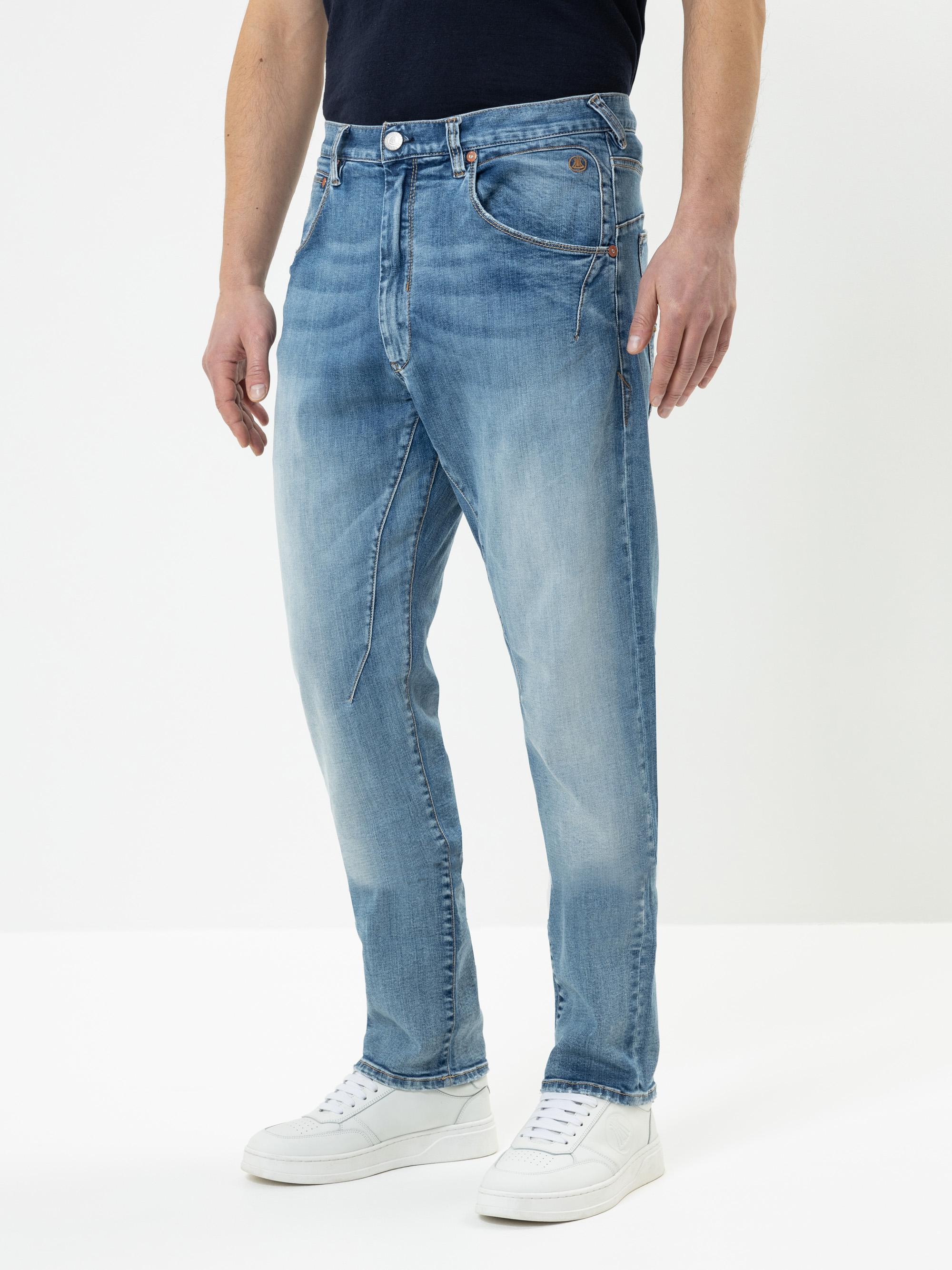 Eine Person in hellblauen Jeans und weißen Turnschuhen steht vor einem einfarbig weißen Hintergrund, wobei nur die untere Hälfte des Körpers sichtbar ist.