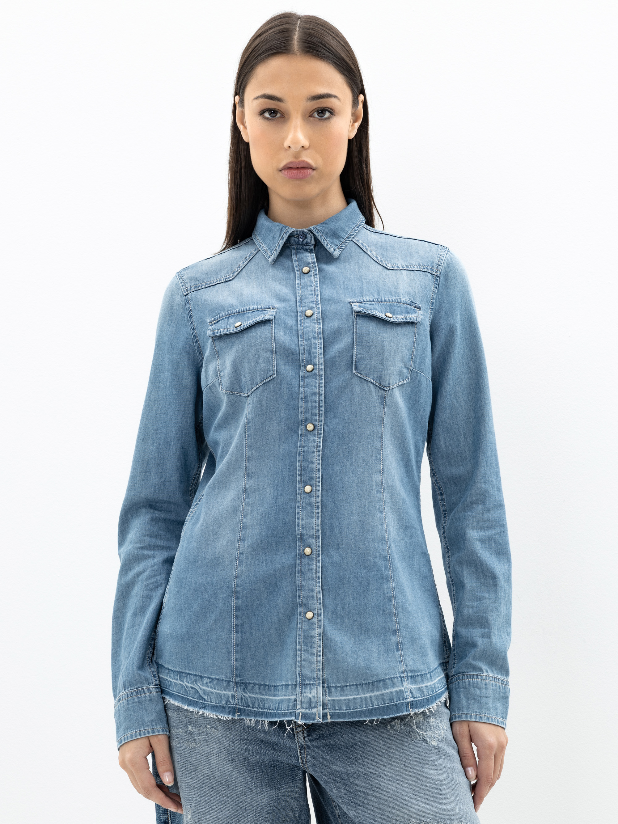 Eine Frau mit glattem braunem Haar trägt ein hellblaues Jeanshemd mit geknöpften Brusttaschen und langen Ärmeln, gepaart mit hellblauen Jeans, vor einem schlichten weißen Hintergrund.